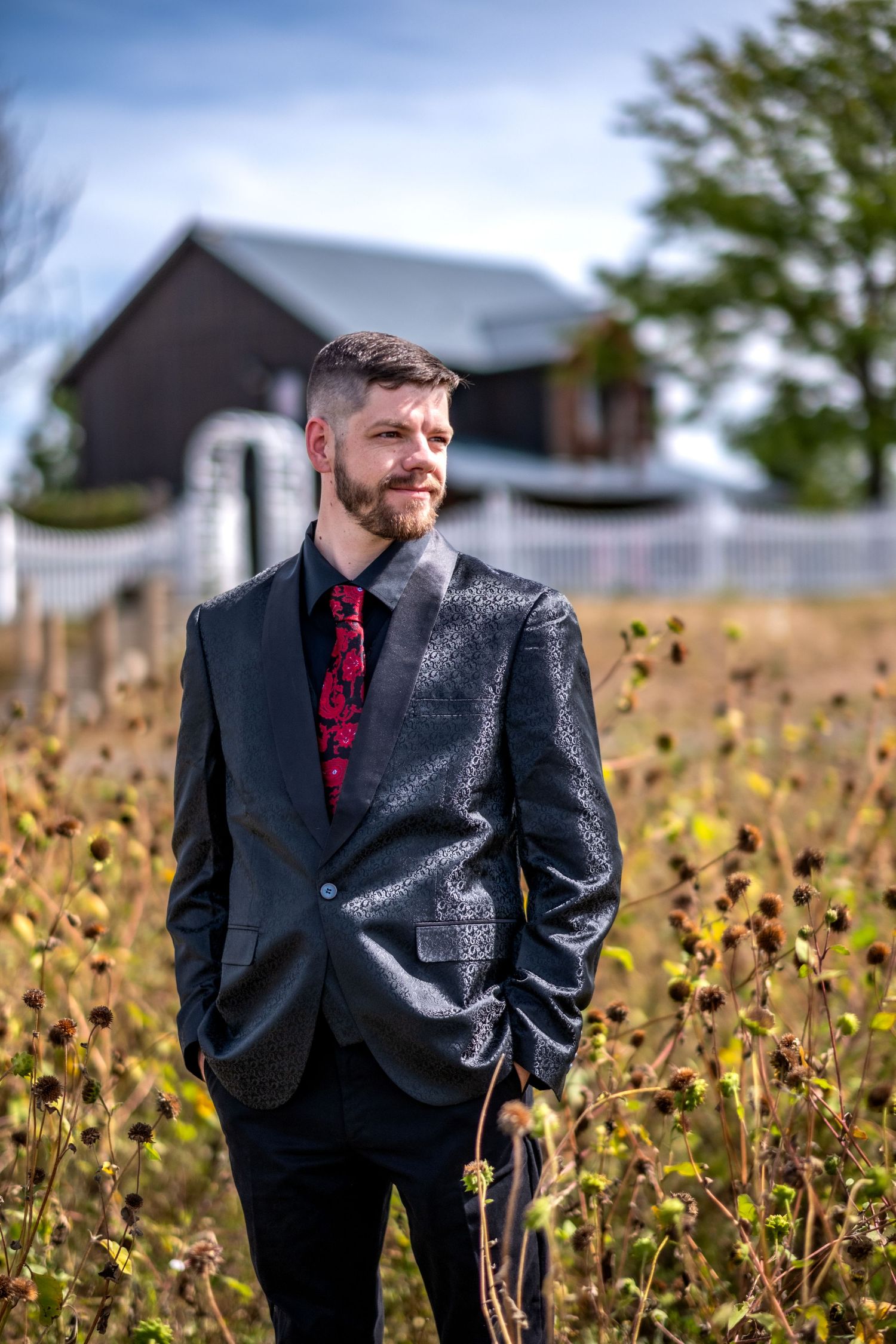 Bearded groom in black suit and red floral tie standing in a field with a barn in the background.