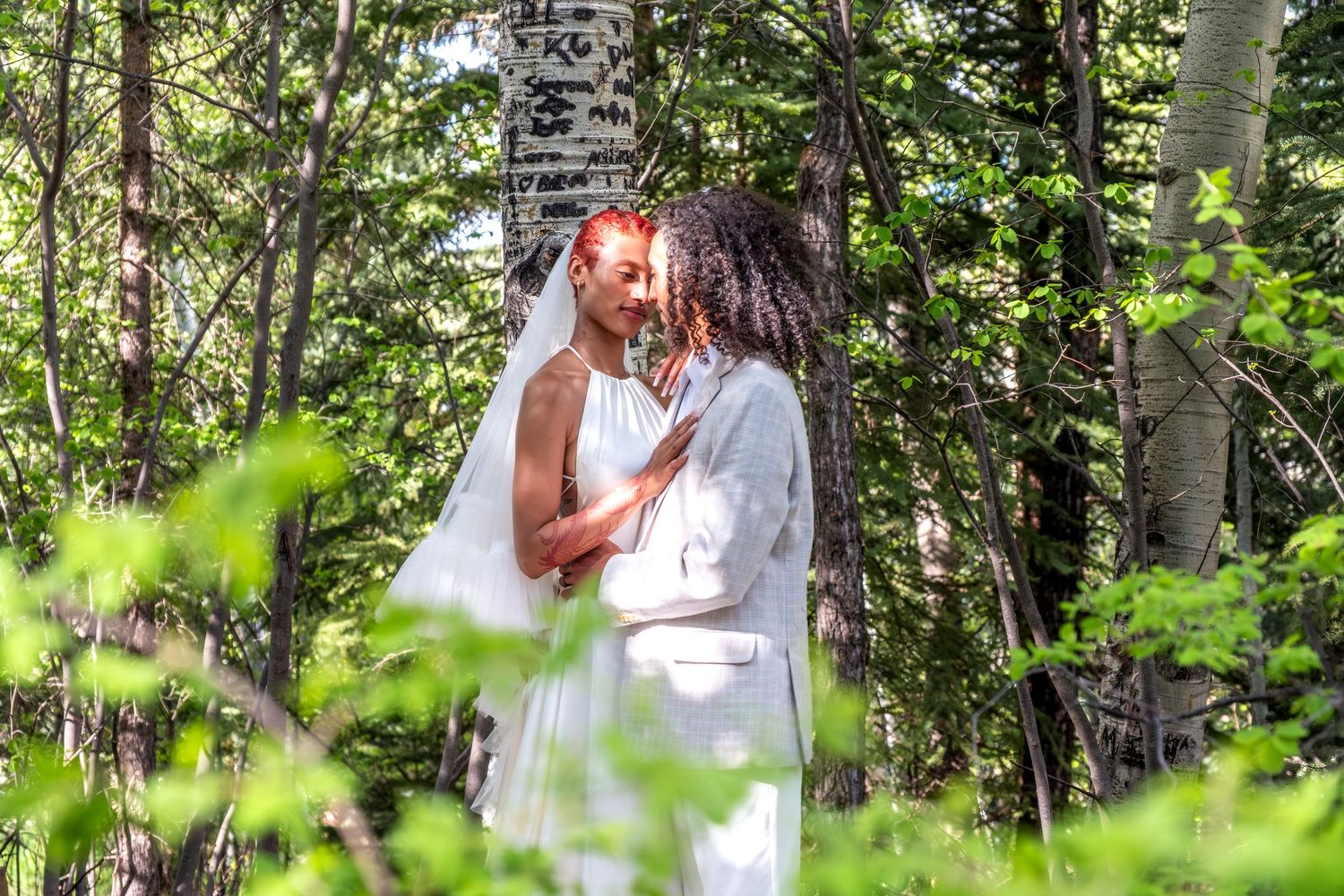 Interracial newlyweds sharing a kiss surrounded by lush green forest trees in outdoor woodland wedding ceremony.