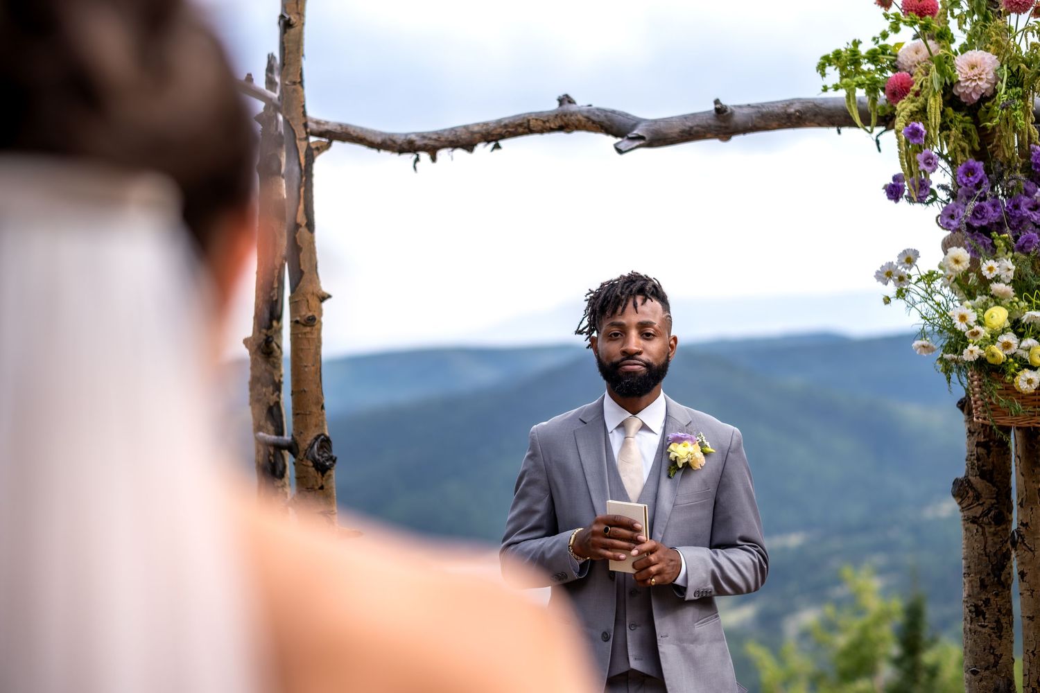 Groom in gray suit stands under rustic floral arch with mountain views during outdoor wedding ceremony.