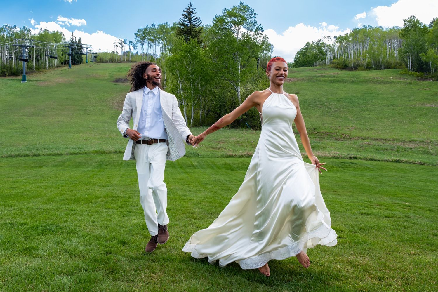 Happy newlywed couple running hand-in-hand across green hillside, bride in flowing white gown, groom in white suit.
