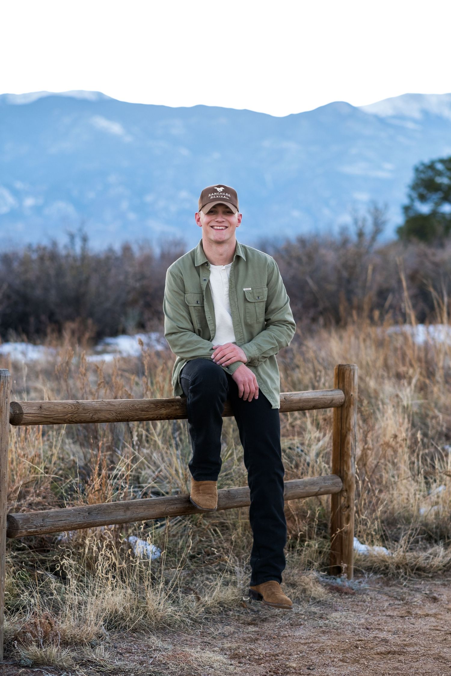 Smiling young man in green shirt and cap sitting on rustic wooden fence with mountain scenery behind.