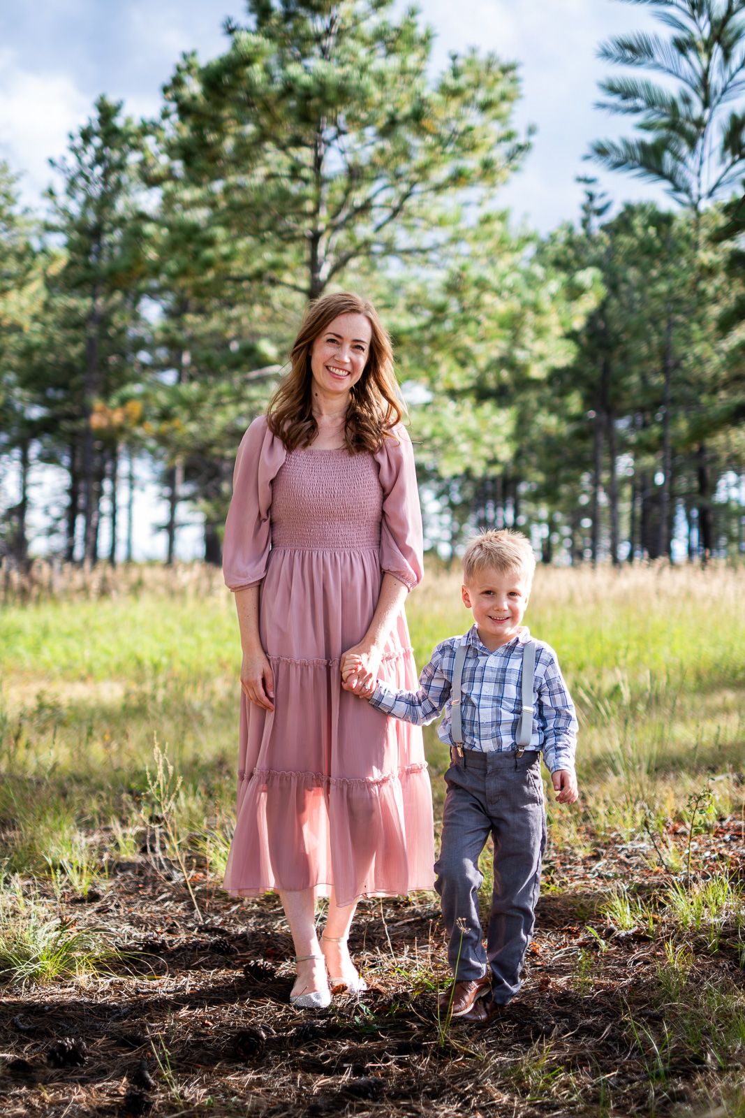 Smiling woman in pink midi dress holds hands with young boy in plaid shirt on a forest trail.