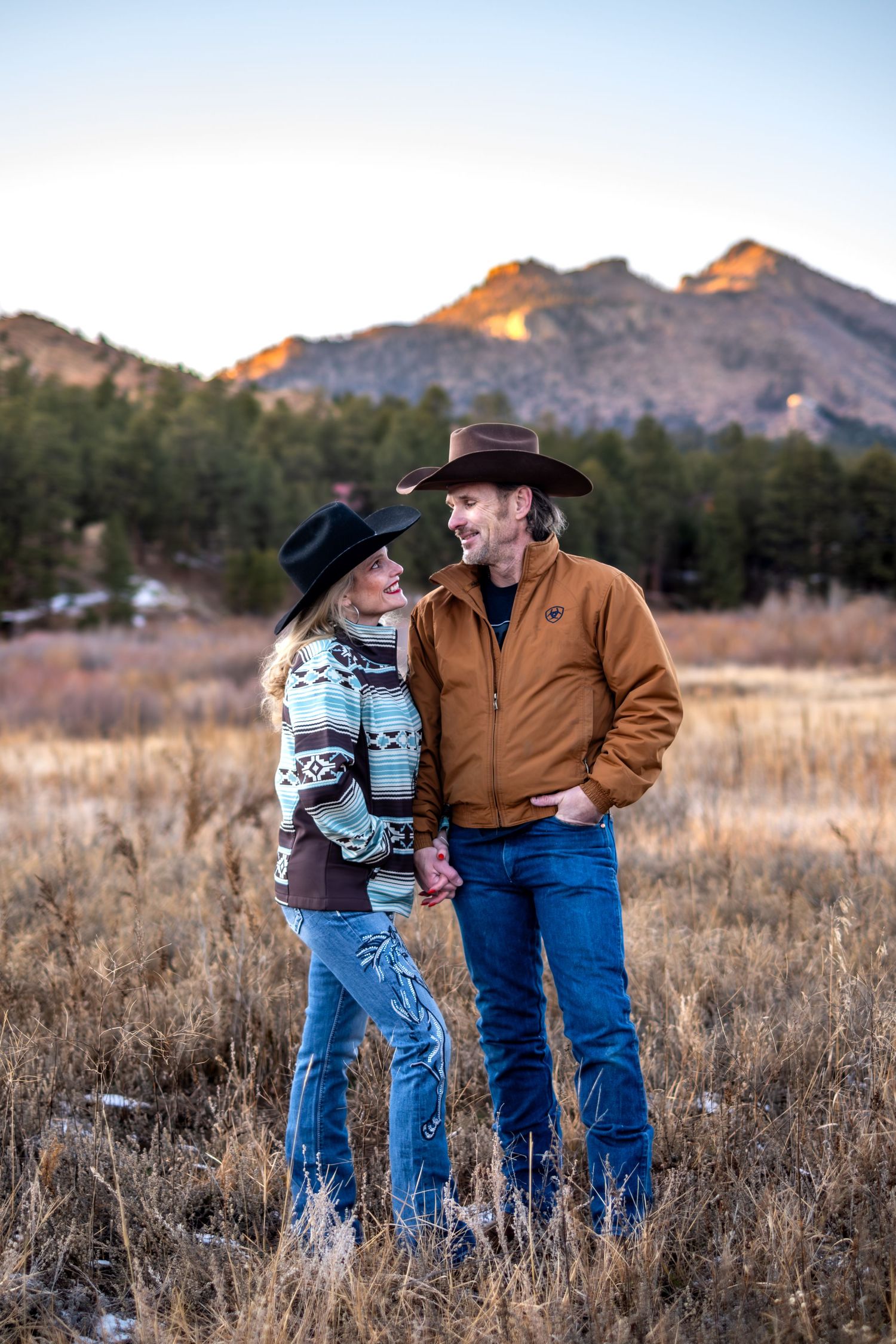 Couple in cowboy hats holding hands in a mountain field at sunset.