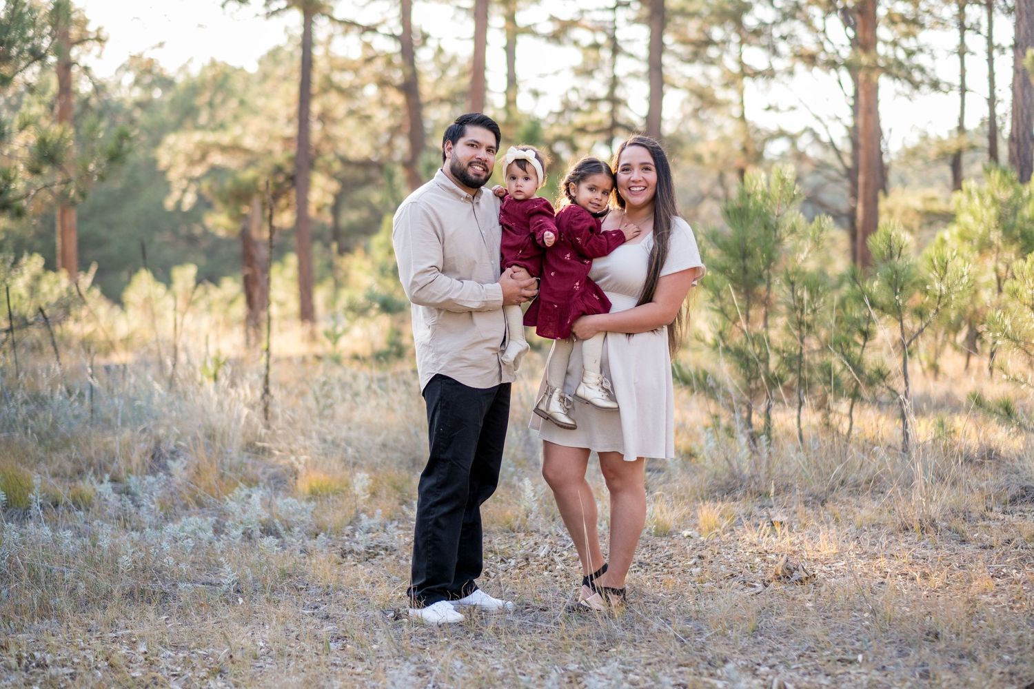 Family of three posing in a pine forest, parents holding toddler in burgundy dress during fall outdoor photo session.