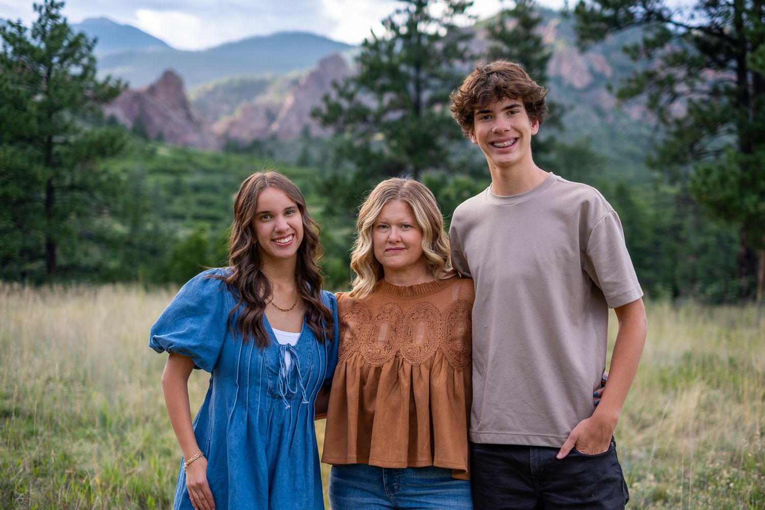 Three teenage siblings smiling together in a meadow with rocky mountain backdrop.