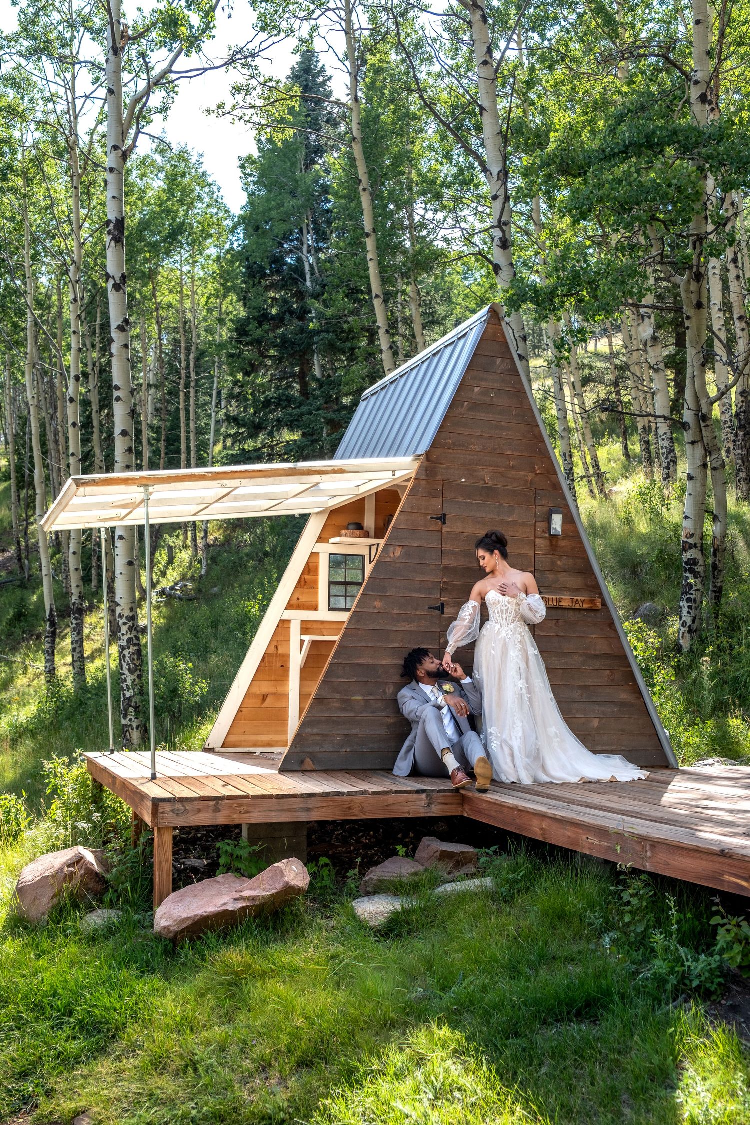 Bride in white gown and groom sitting on the deck of a rustic A-frame cabin in an aspen forest.