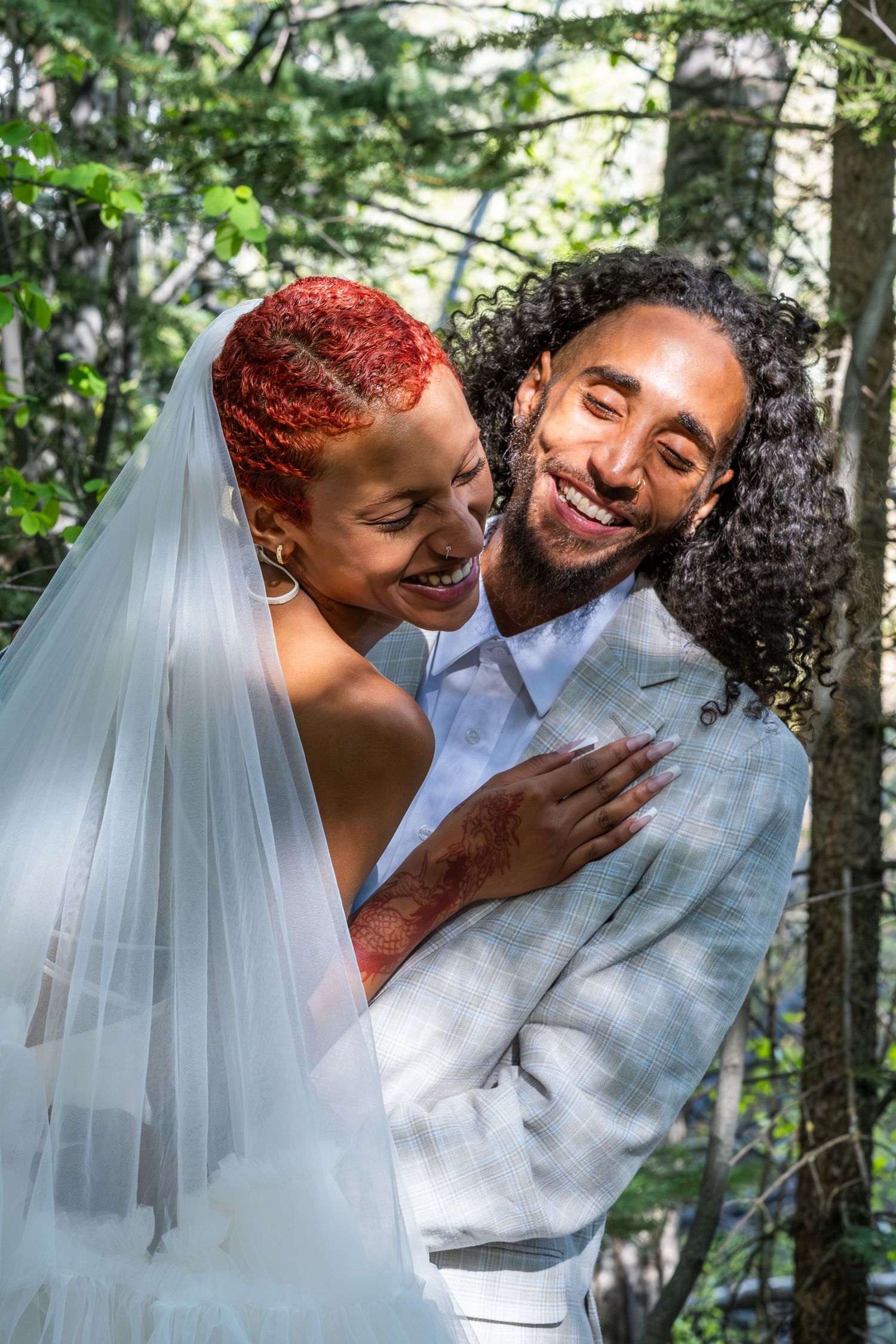 Joyful interracial couple embracing in forest wedding attire, bride in white veil with red hair, groom in gray suit smiling.