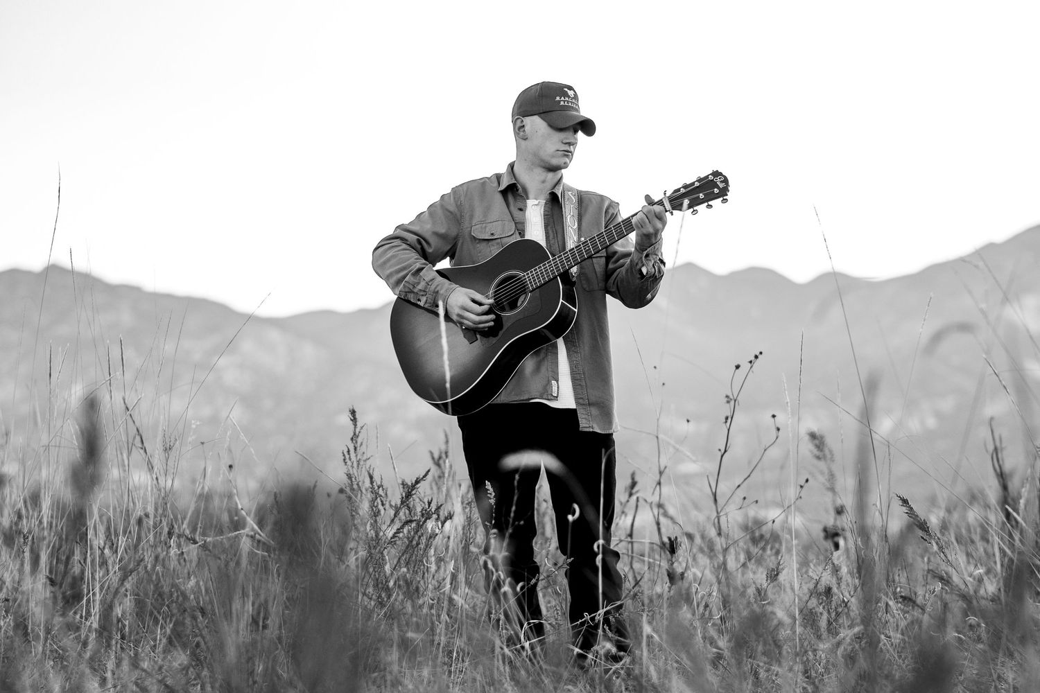 Black and white photo of young male musician playing acoustic guitar in open field with mountains.