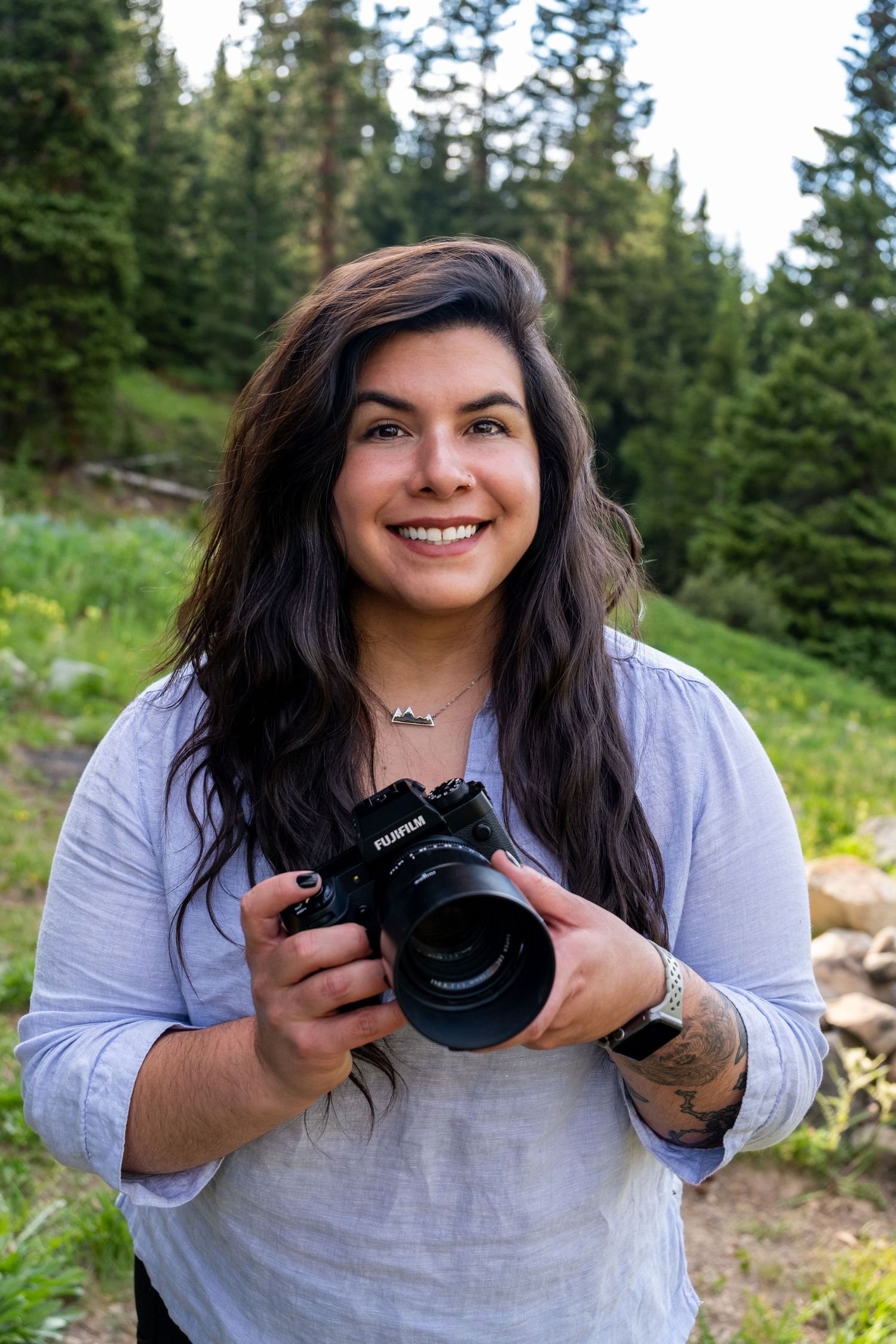 Smiling woman holding a Fujifilm camera outdoors in a lush mountain forest setting.