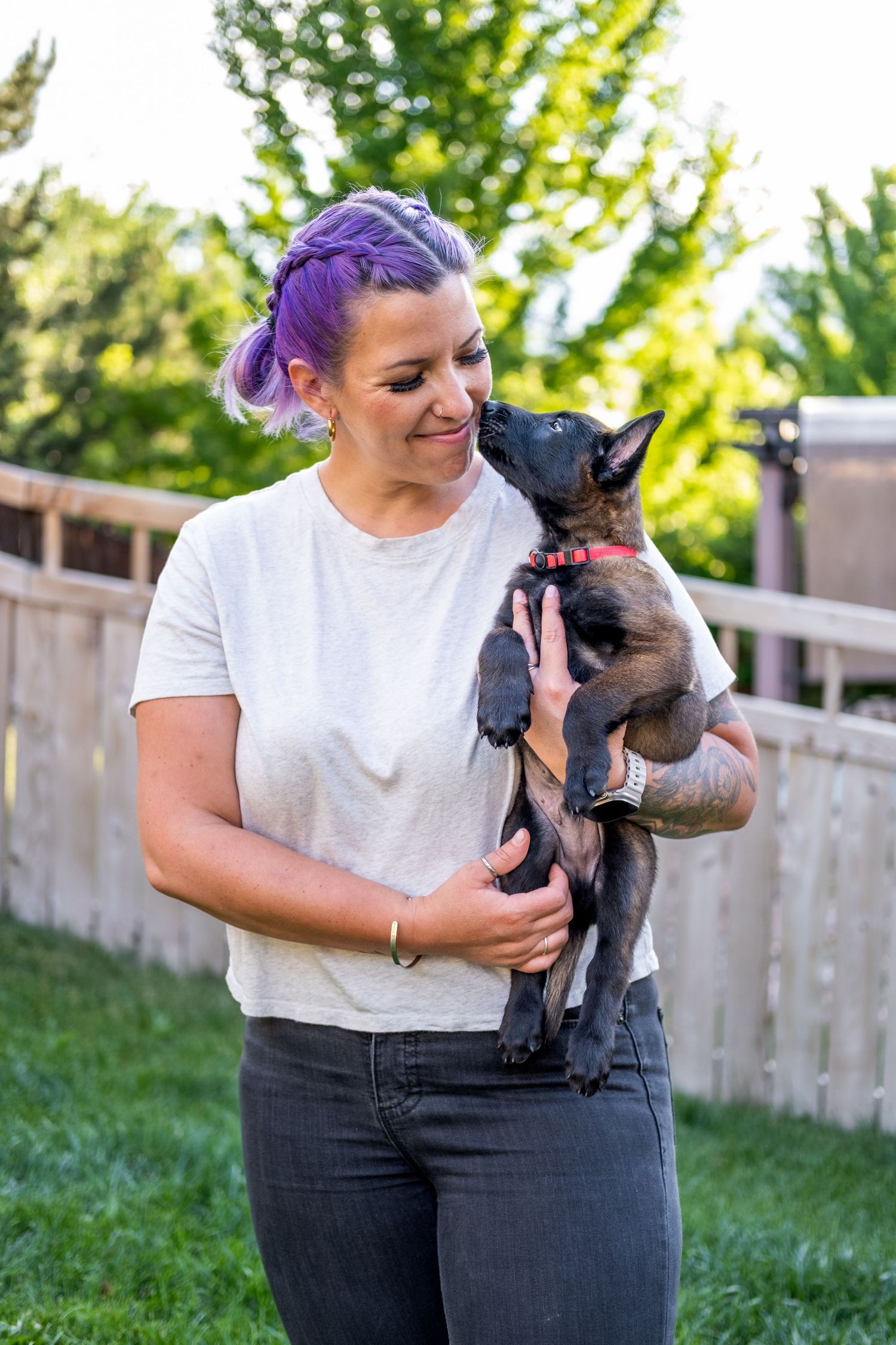 Woman with purple hair smiling while holding a small black puppy with a red collar outdoors.