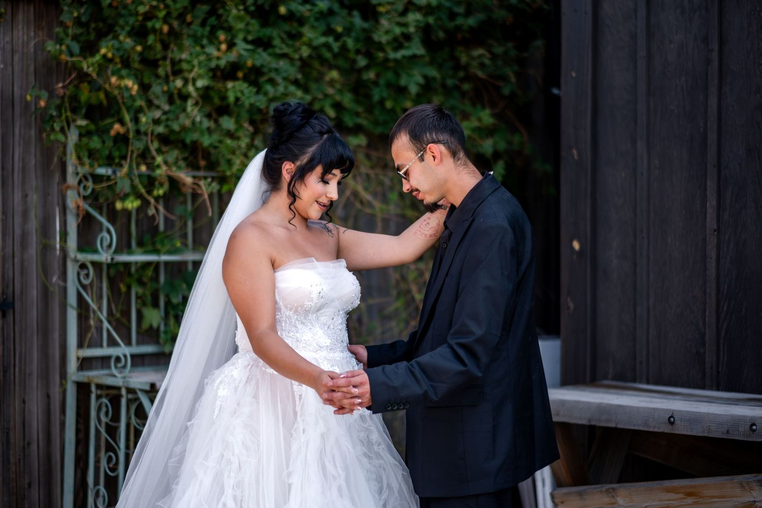 Bride in white strapless gown holding hands with groom in black suit near a wooden structure.