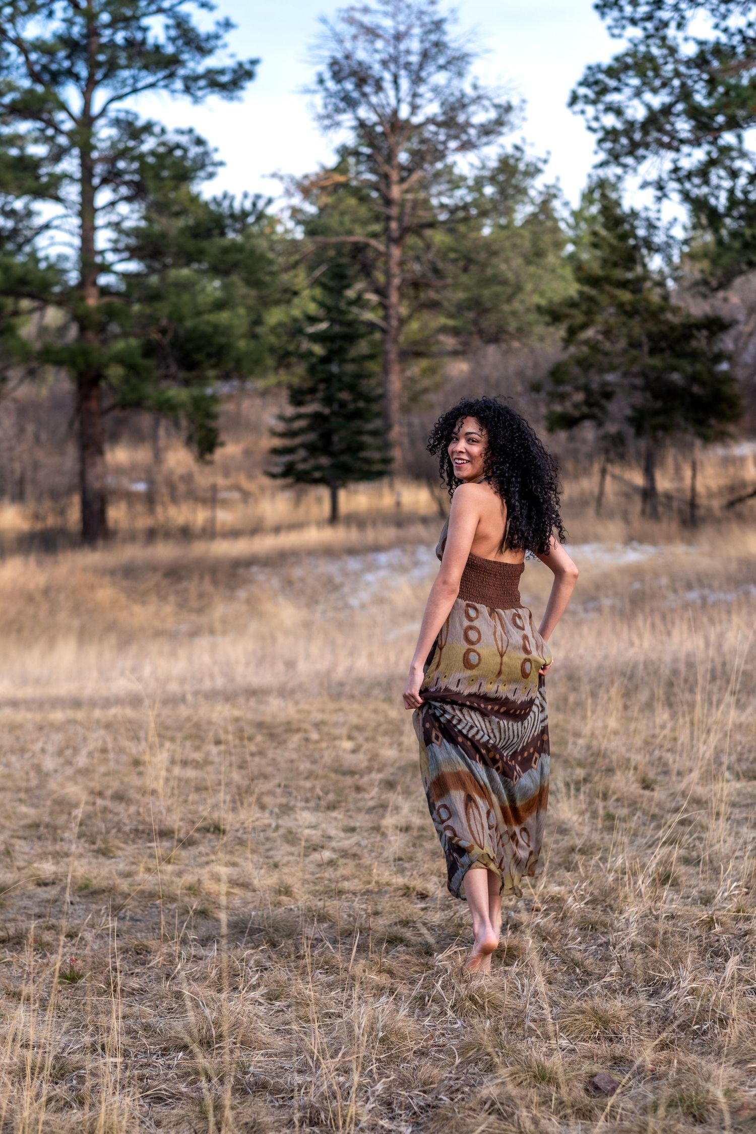 Smiling woman in bohemian dress walking barefoot through dry grass field with pine trees.