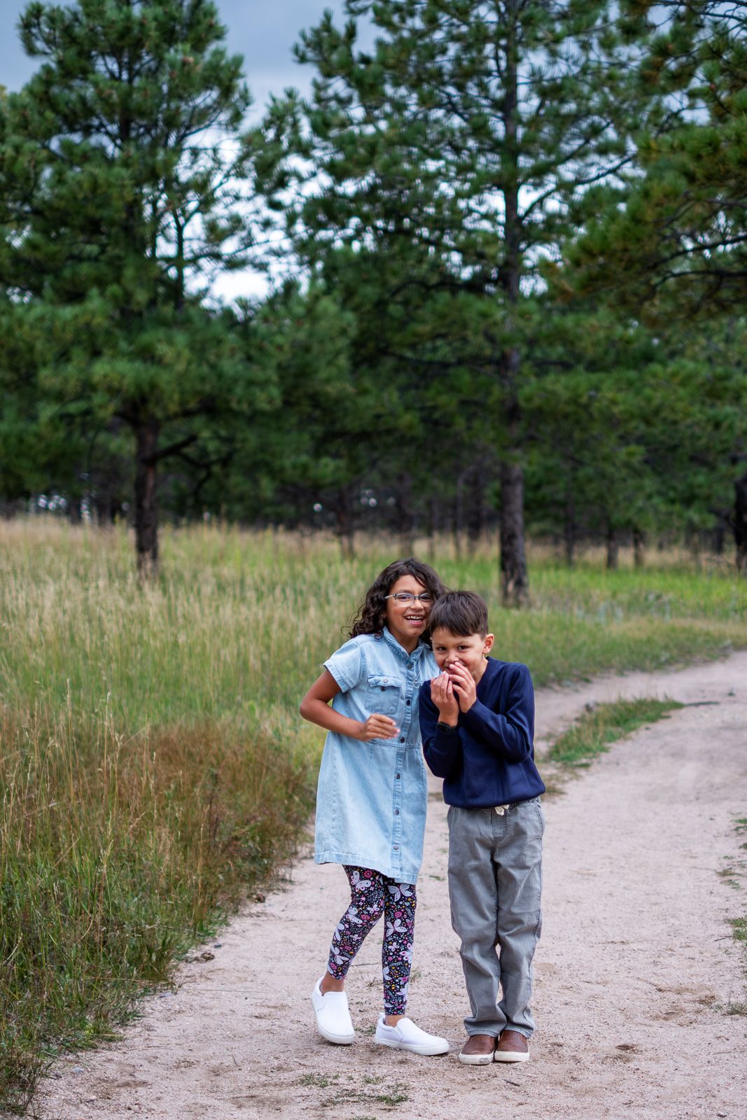 Two siblings laughing and playing together on a sandy forest trail surrounded by tall green pine trees.