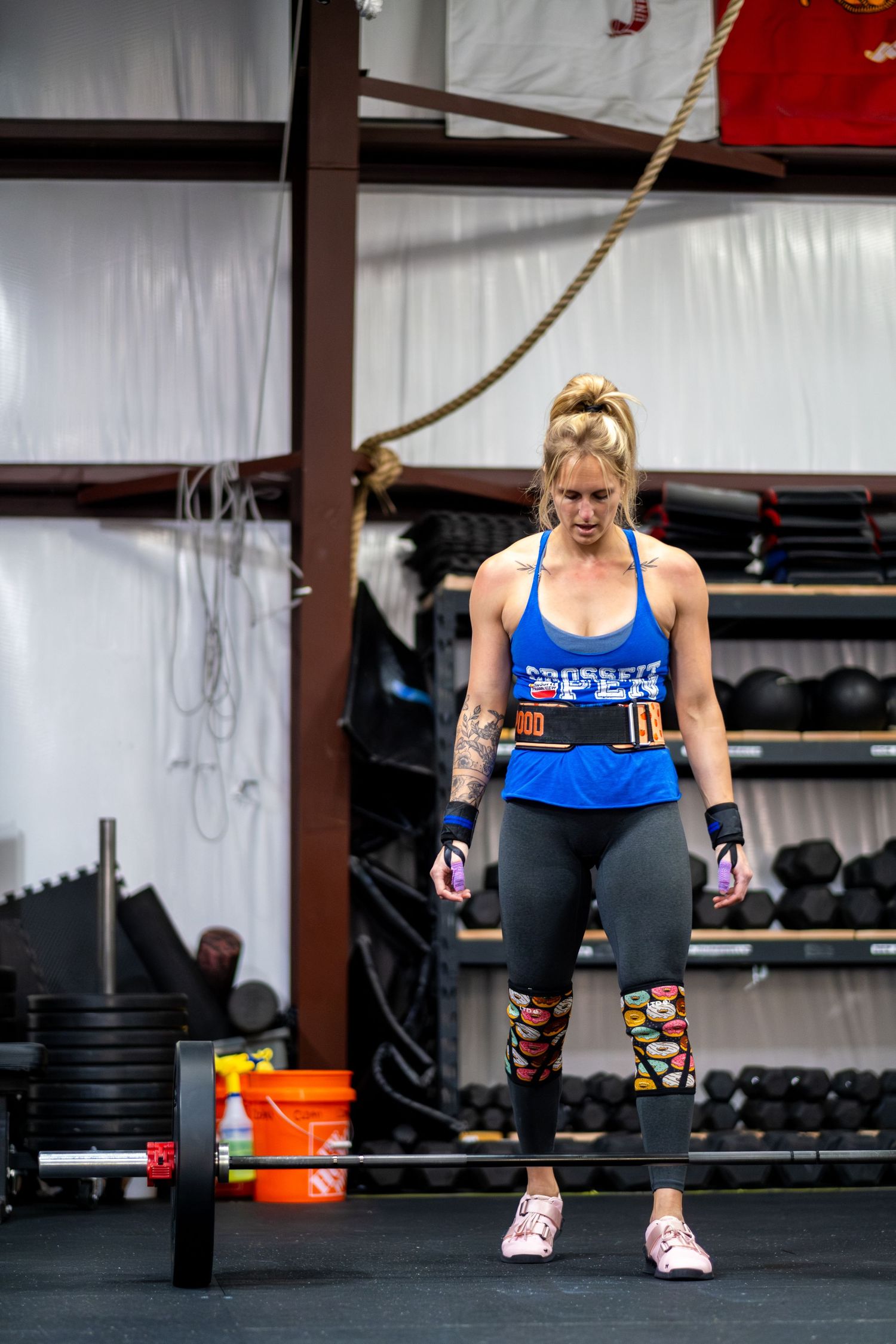 Woman in blue tank top prepares to deadlift a barbell in a CrossFit gym wearing a weightlifting belt.