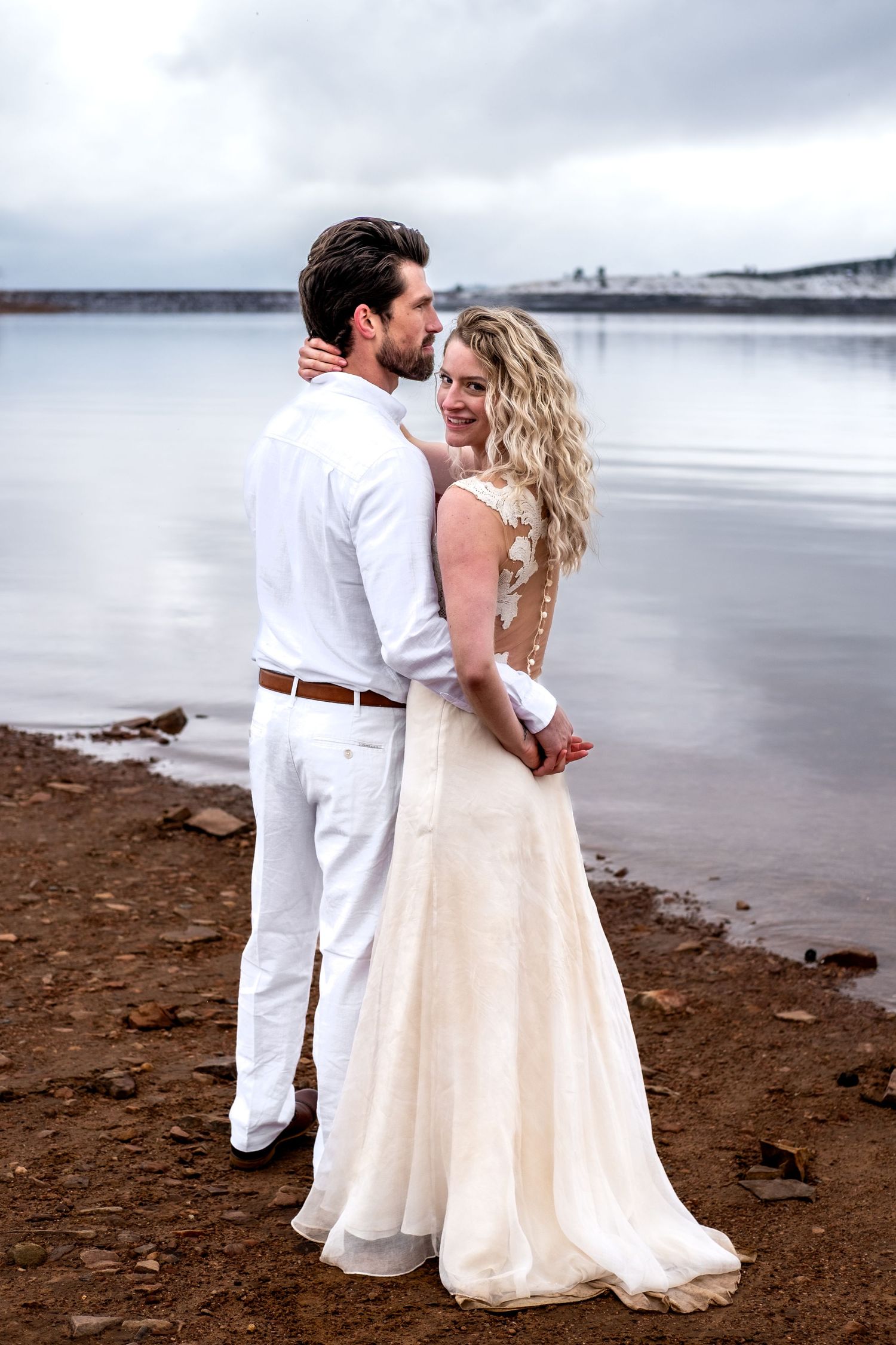 A couple in wedding attire embraces by a lakeside, the bride in a white gown smiling over her shoulder.
