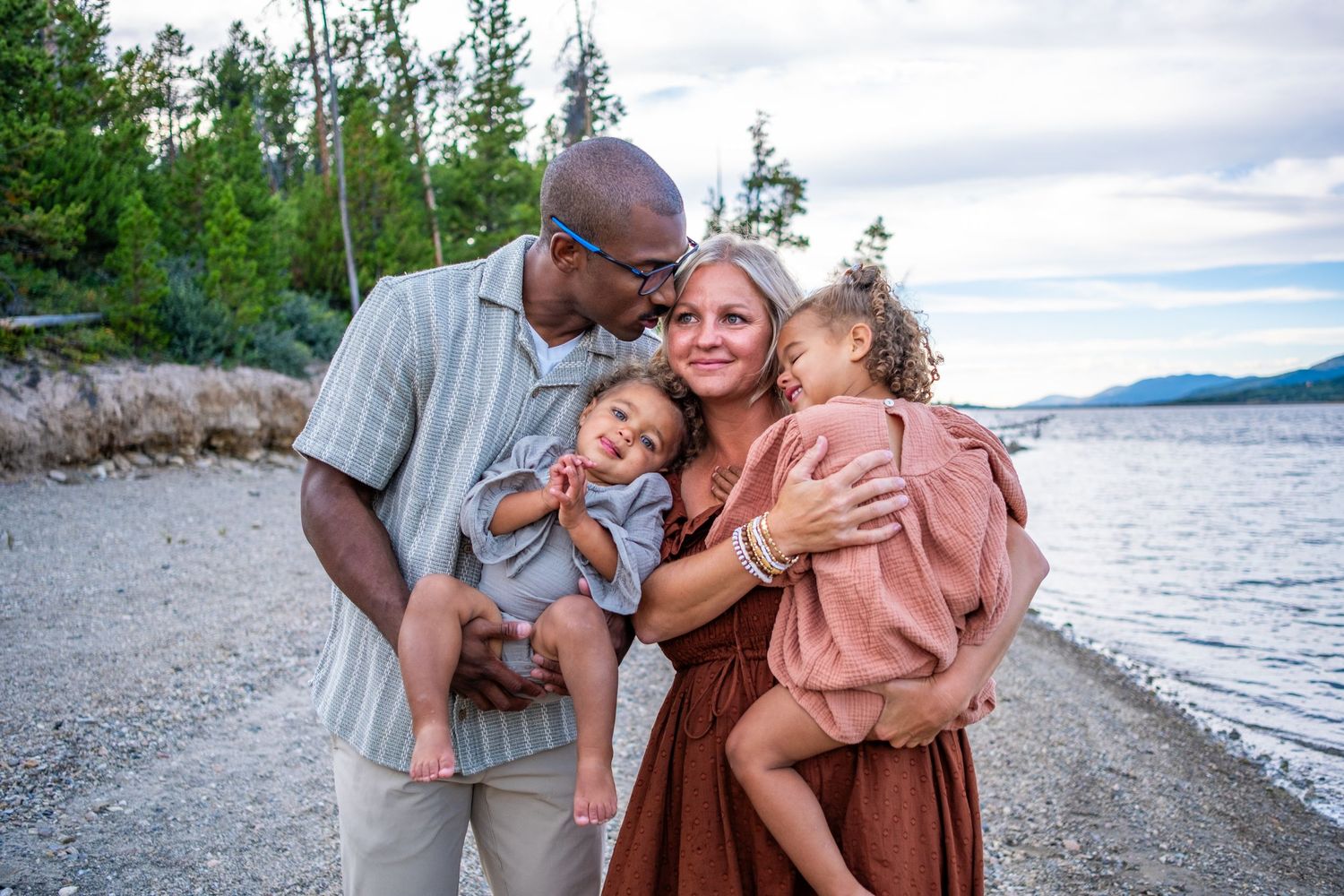 Loving interracial family with two young children sharing a tender moment on a scenic lakeside beach.