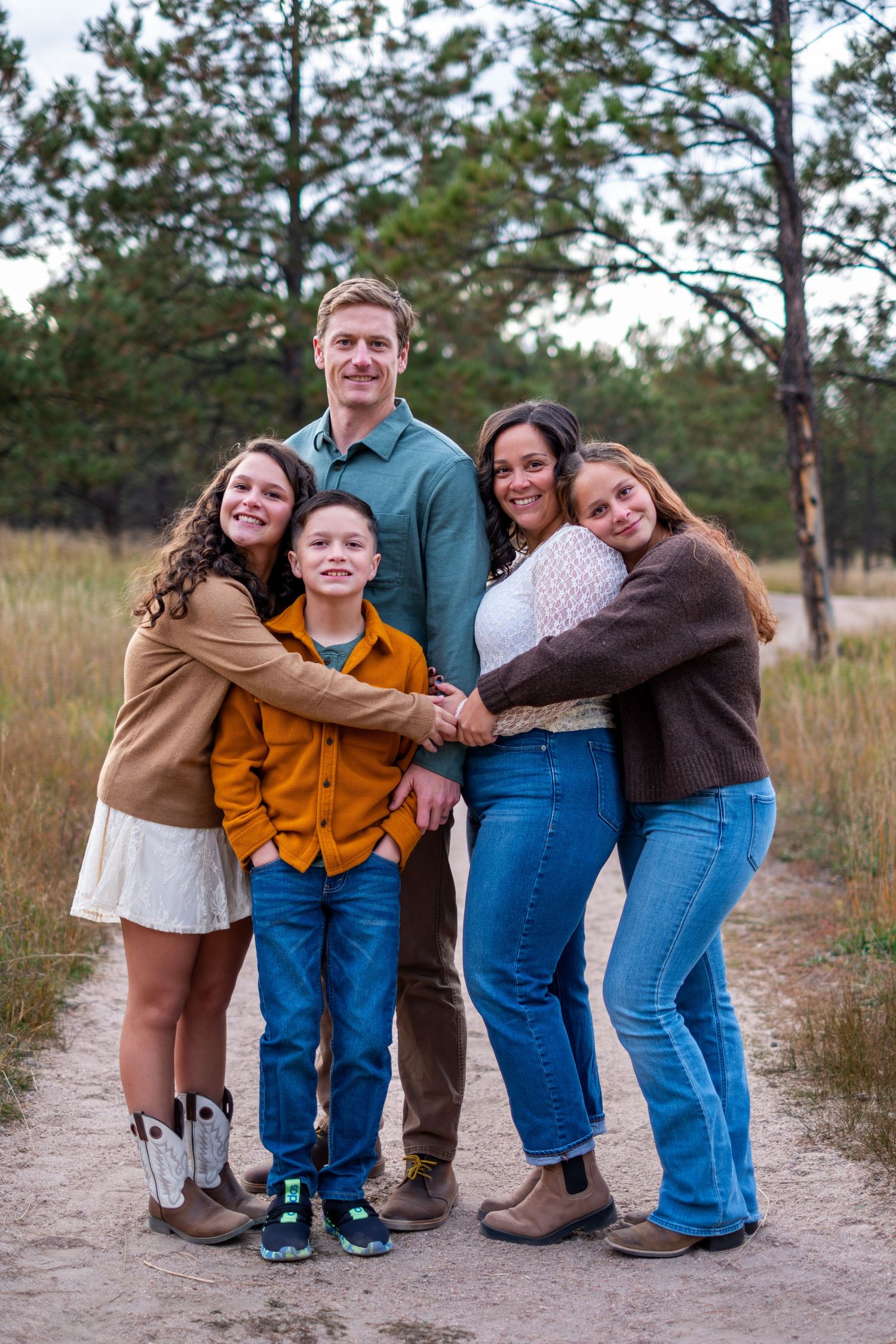 Happy family of five hugging together on a dirt path surrounded by pine trees and tall grass.