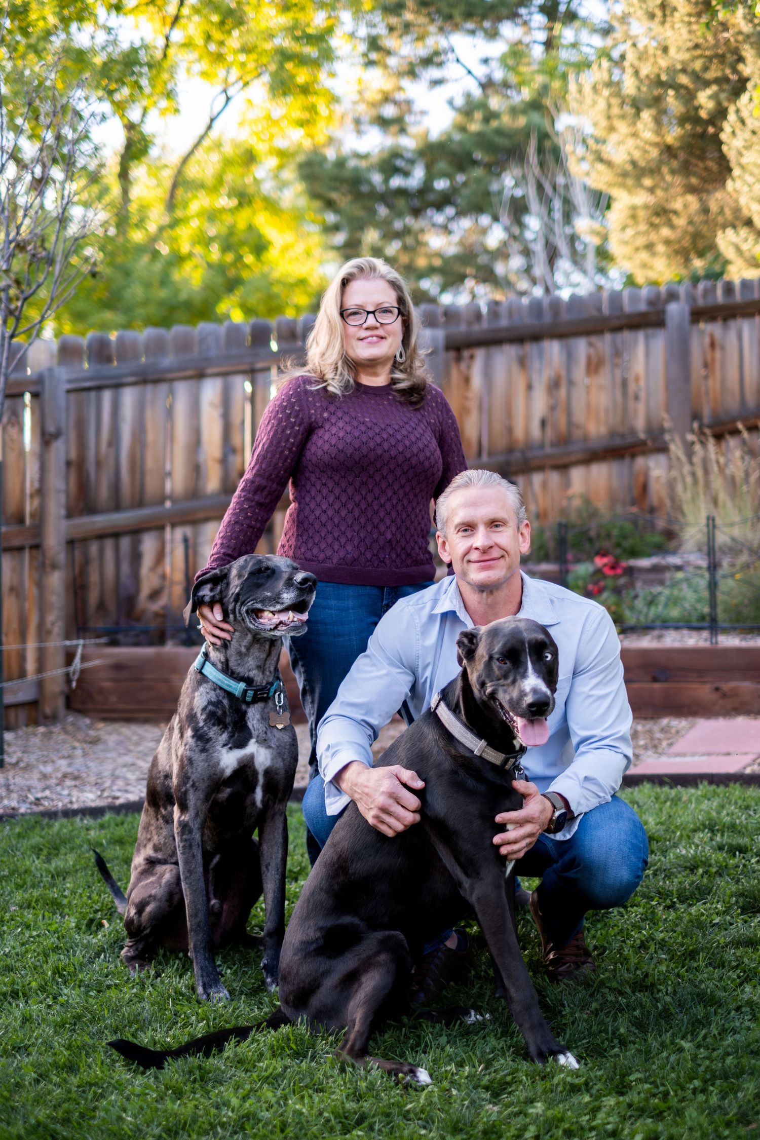 Couple poses in backyard with two large black dogs on green grass near wooden fence.