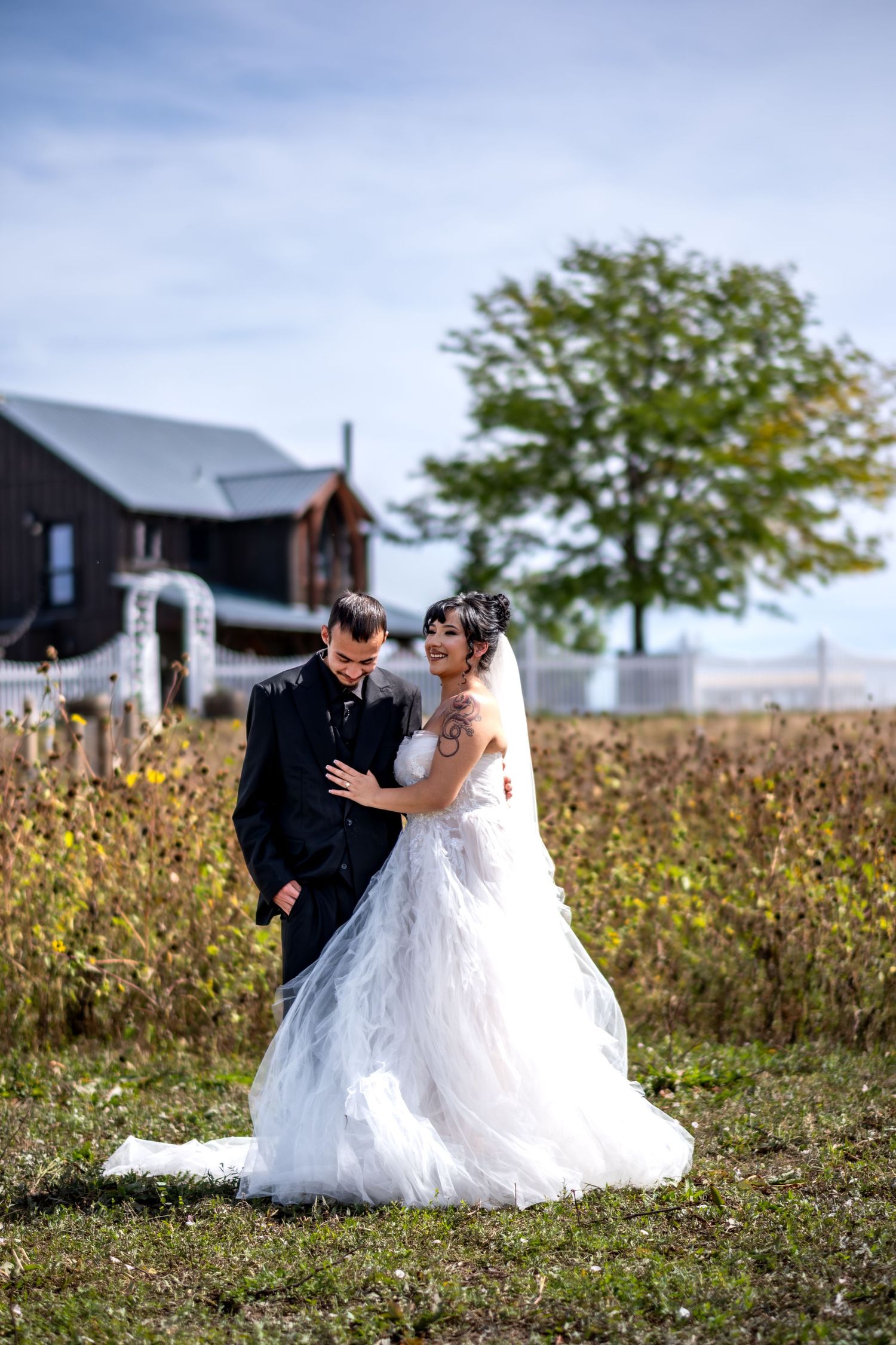 Bride in white tulle gown and groom in black suit embracing in a field with a barn backdrop.