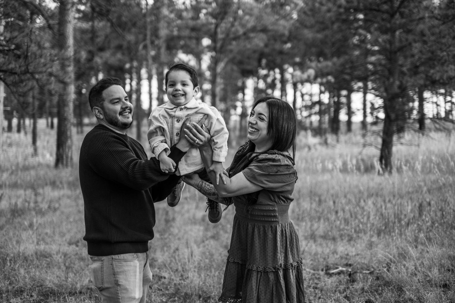 Black and white photo of parents joyfully lifting their toddler in a pine forest meadow.