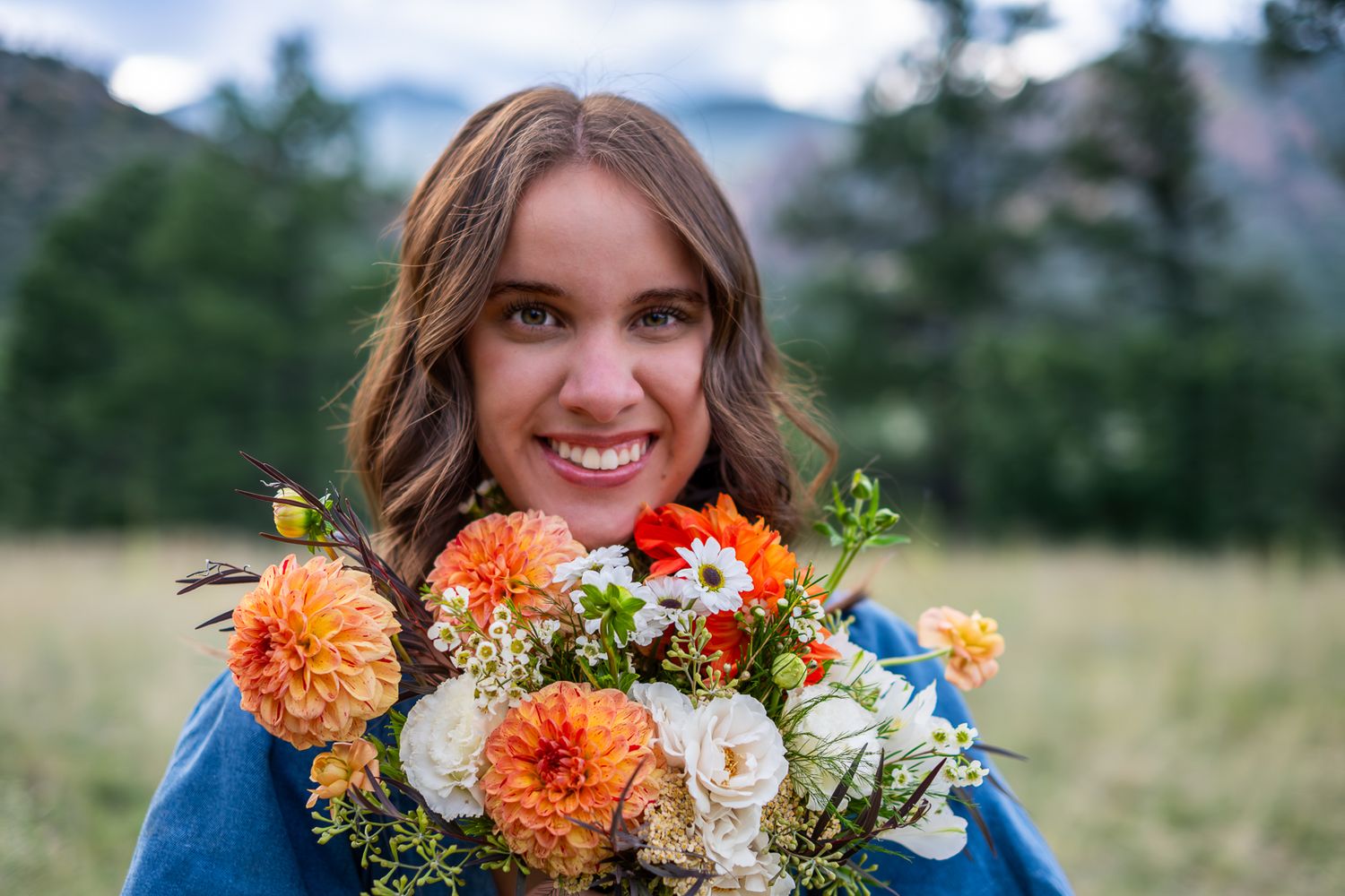 Smiling woman holding vibrant orange and white flower bouquet with mountains in background.