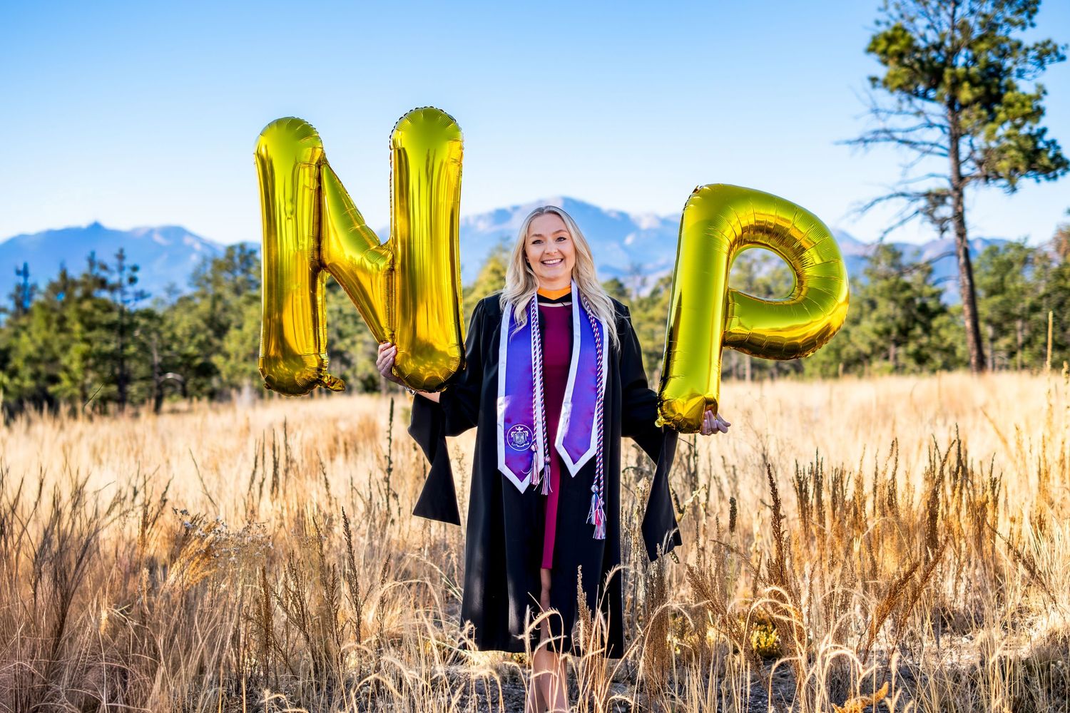 Blonde woman in graduation gown holding gold NP balloons in golden mountain field.