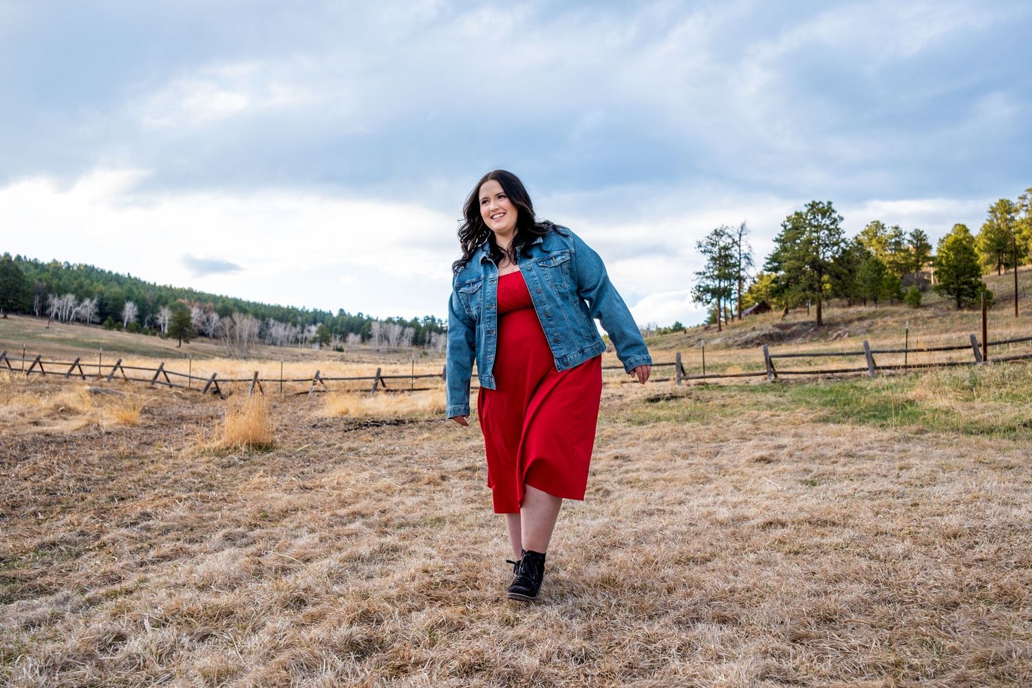 Young woman in red dress and denim jacket walking through an open field under a dramatic cloudy sky.