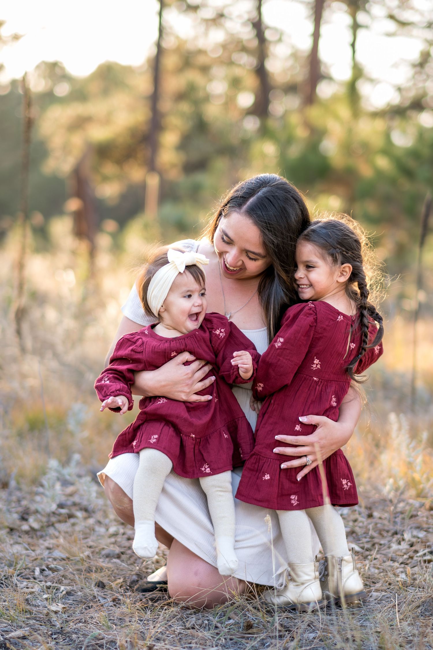 Mother kneeling outdoors hugging two young daughters in matching burgundy dresses in a sunlit pine forest.
