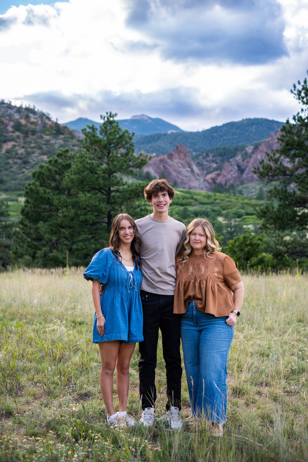 Three young people posing together in a grassy mountain meadow with pine trees and red rock formations.