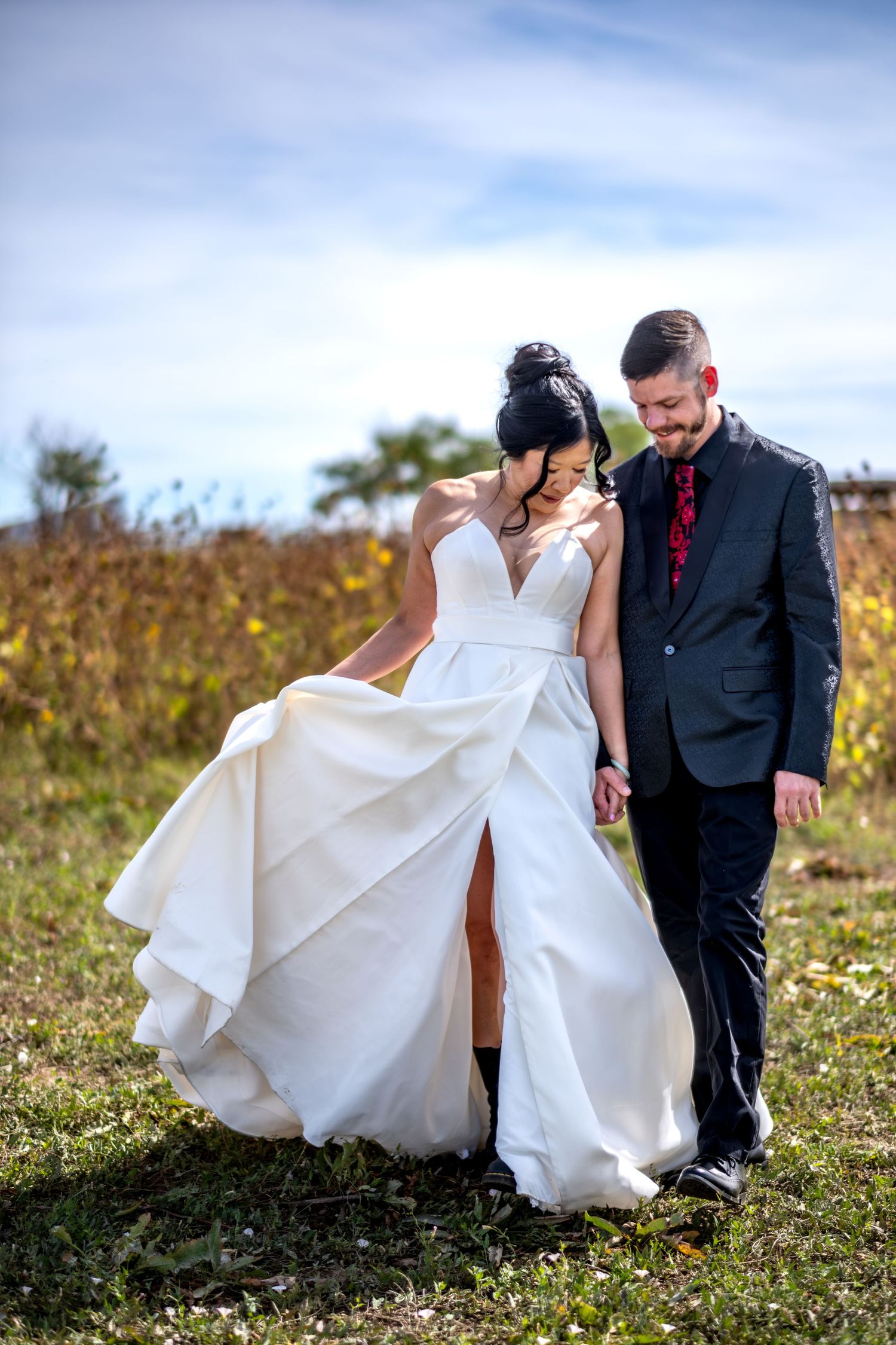 A bride in a white ballgown and groom in a dark suit walk hand in hand through an autumn field.