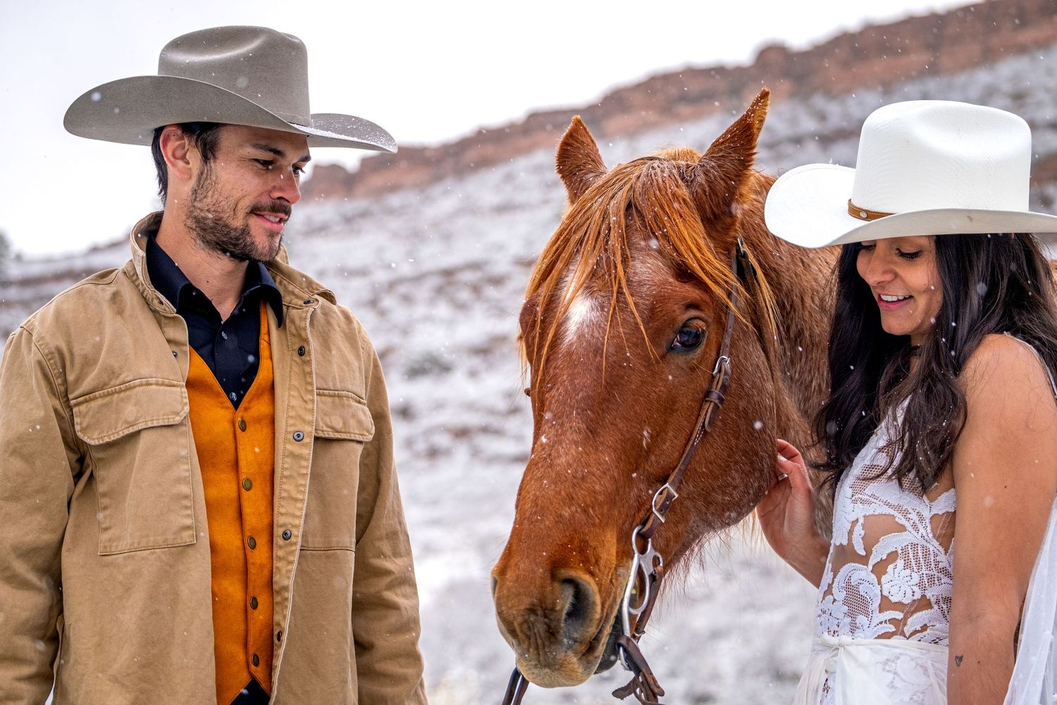 Smiling western wedding couple in cowboy hats standing closely with chestnut horse in snowy desert.