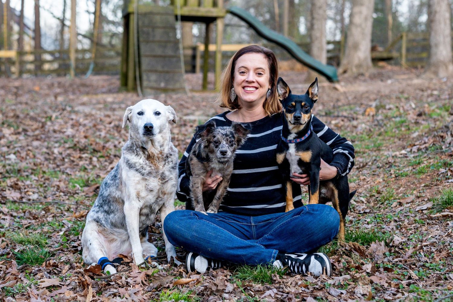 Smiling woman sitting cross-legged outdoors with three dogs of different breeds beside her.