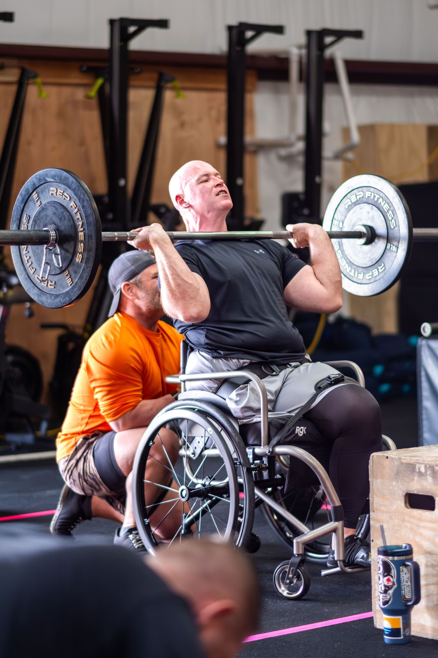 Adaptive athlete in a wheelchair performing a barbell back squat with a spotter at a CrossFit gym.