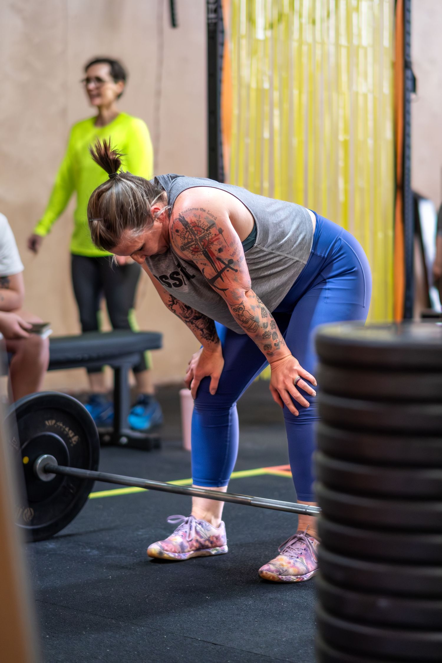 Tattooed woman in blue leggings prepares to deadlift a barbell at a CrossFit gym competition.