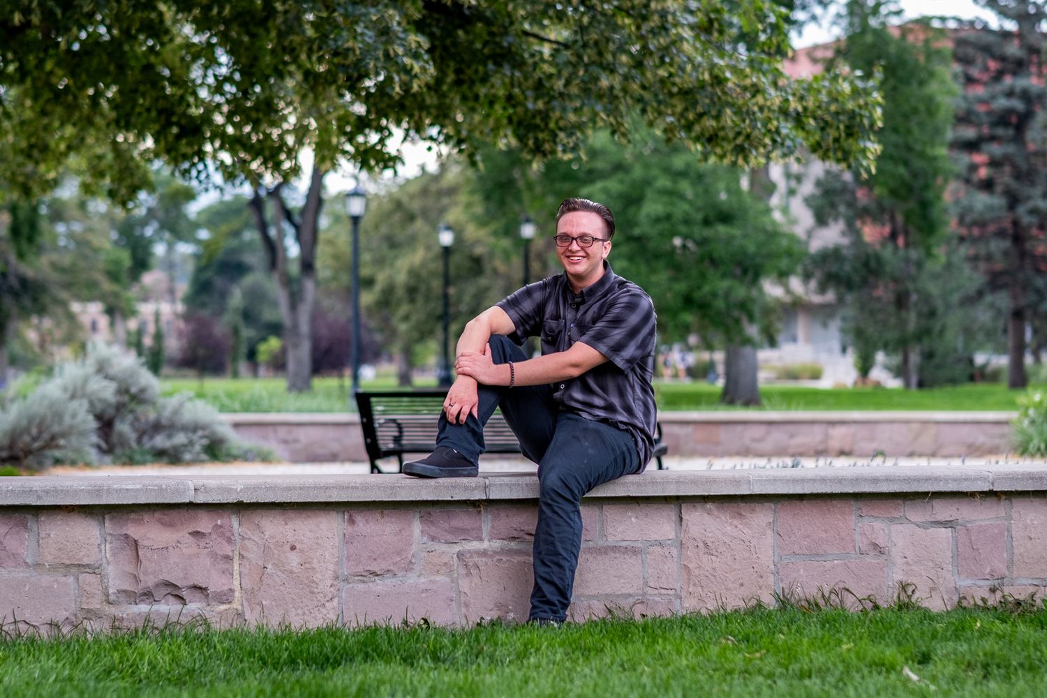 Smiling man with glasses sitting on a stone wall with a laptop in a lush green campus park.