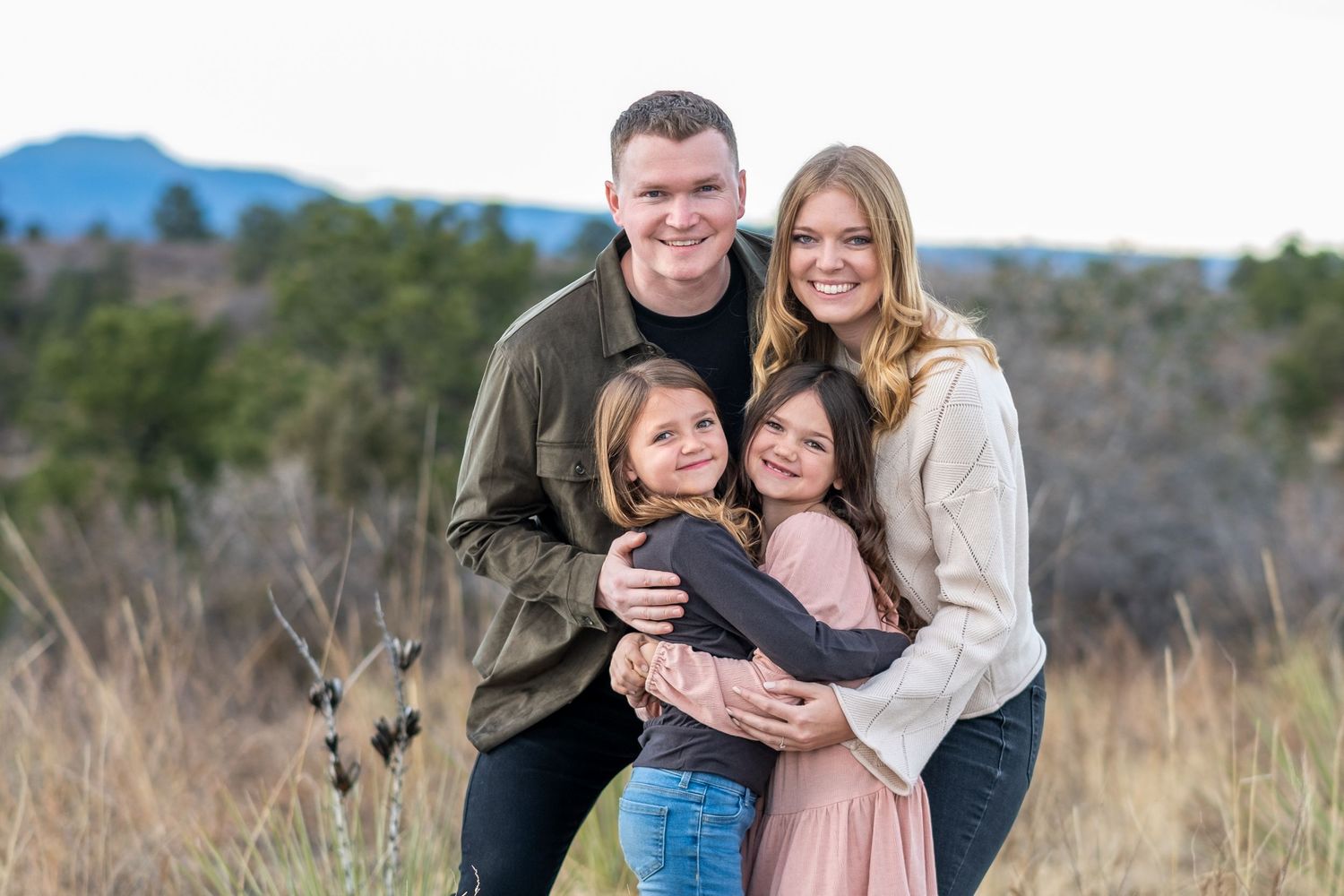 Happy family of four embracing in golden field, parents with two daughters during outdoor fall photo session.