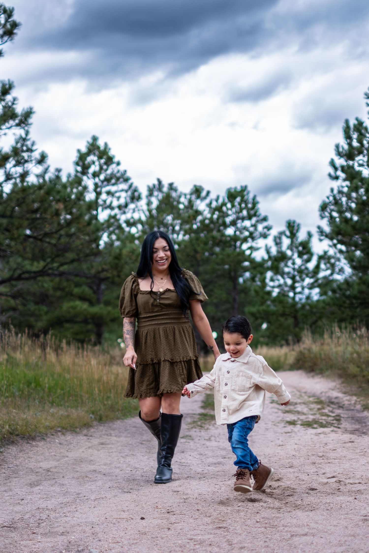 Mother in olive dress and boots walking hand in hand with young son on forest path.