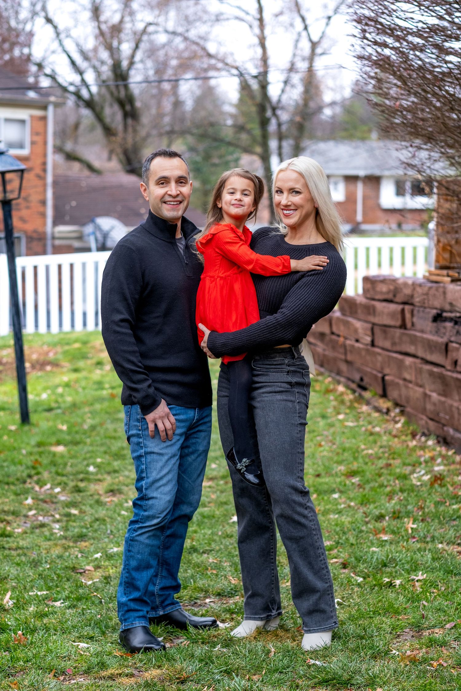 Happy family of three posing outdoors, parents holding young daughter in red dress in suburban yard.