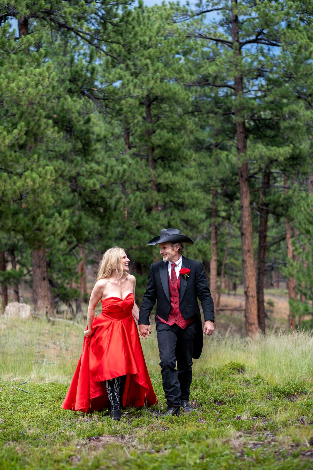 Western wedding couple holding hands walking through pine forest, bride in red gown and groom in black cowboy hat.