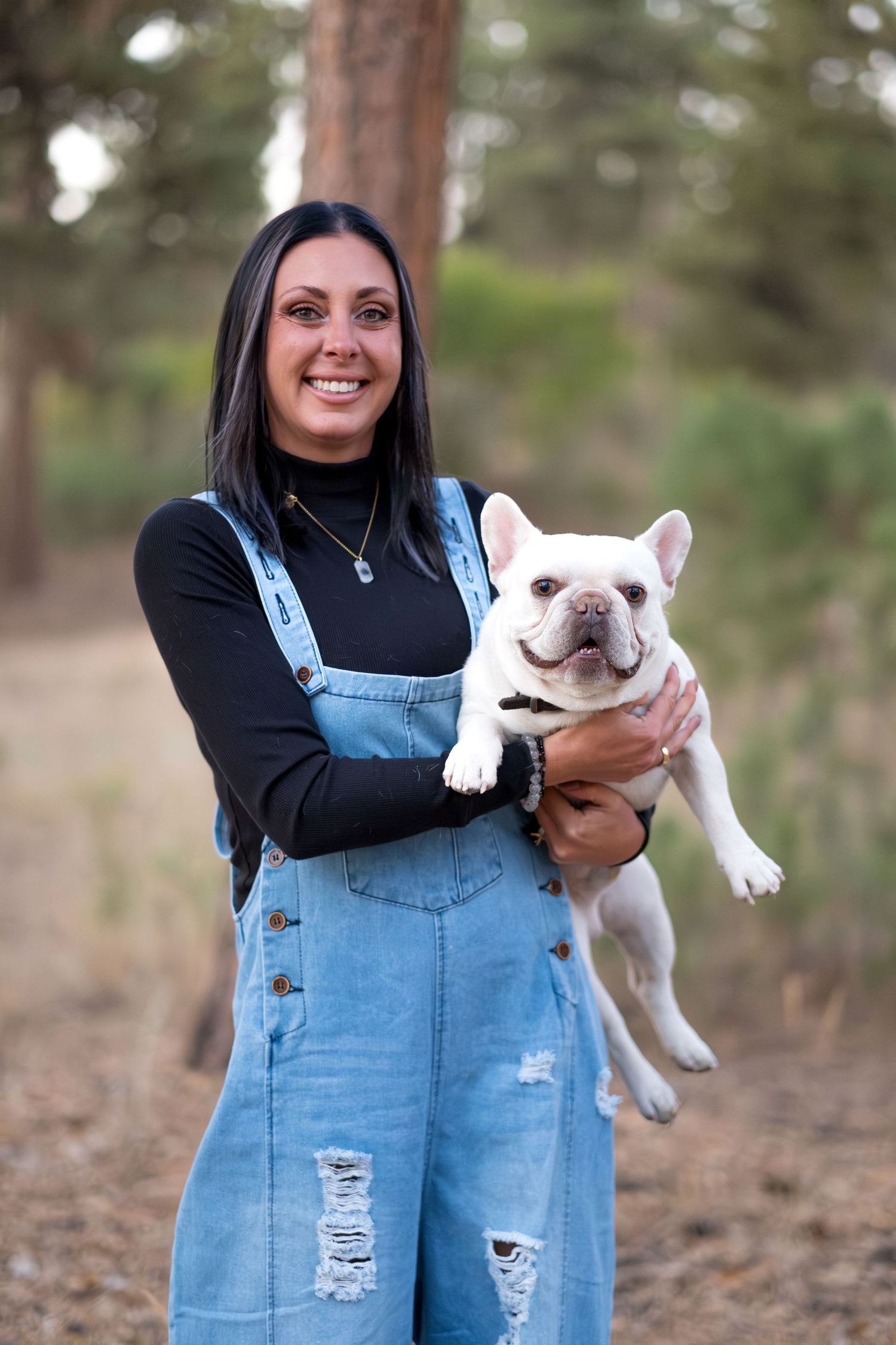 Smiling woman in denim overalls holding a white French Bulldog in a pine forest path.