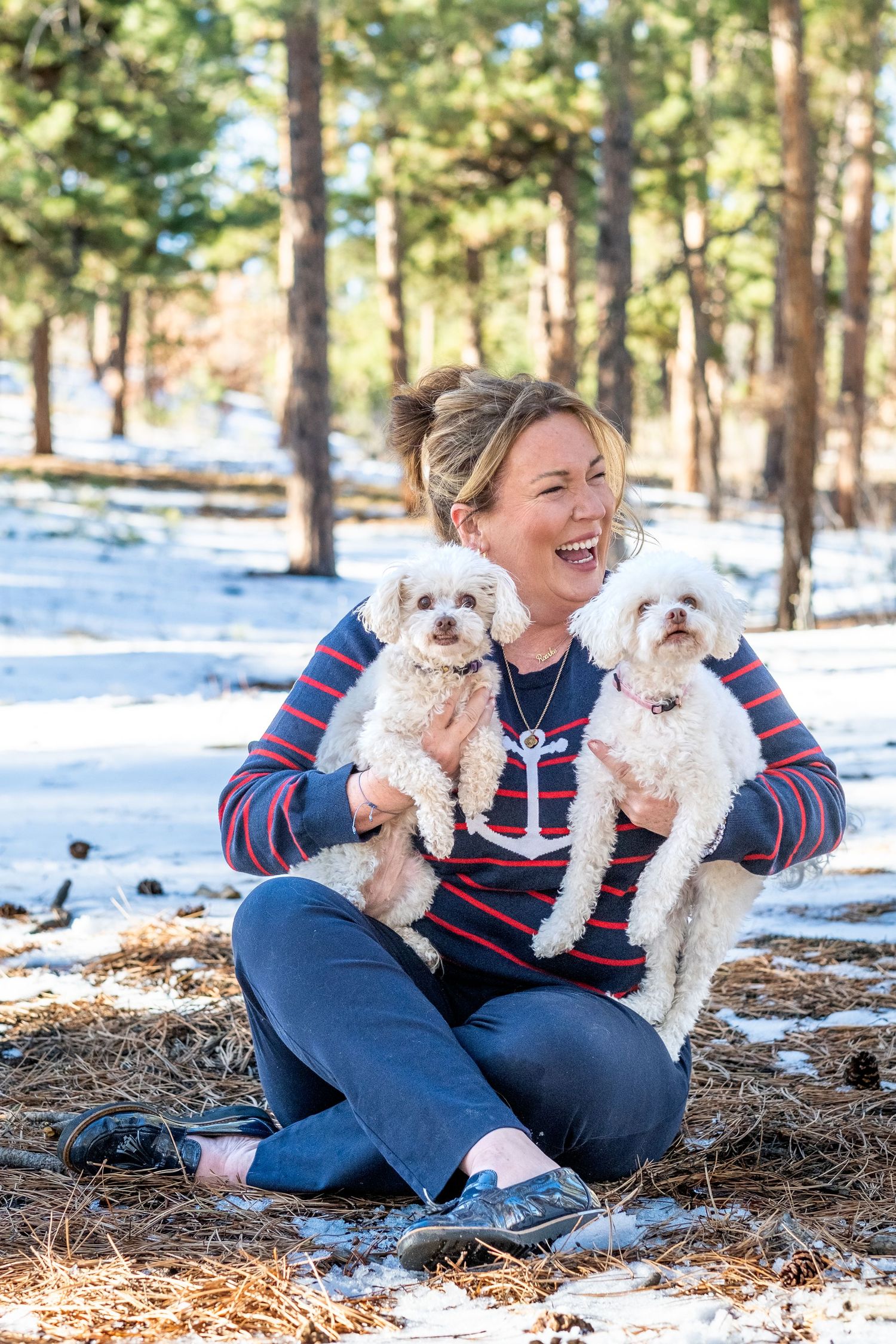 Laughing woman sitting in snowy pine forest holding two fluffy white Bichon Frise dogs.