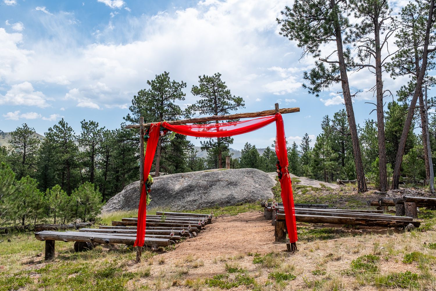Rustic outdoor wedding arch decorated with red fabric draping between pine trees with wooden bench seating.