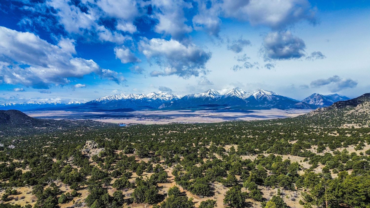 Panoramic aerial view of a vast valley with snow-capped mountain peaks under dramatic blue skies and white clouds.