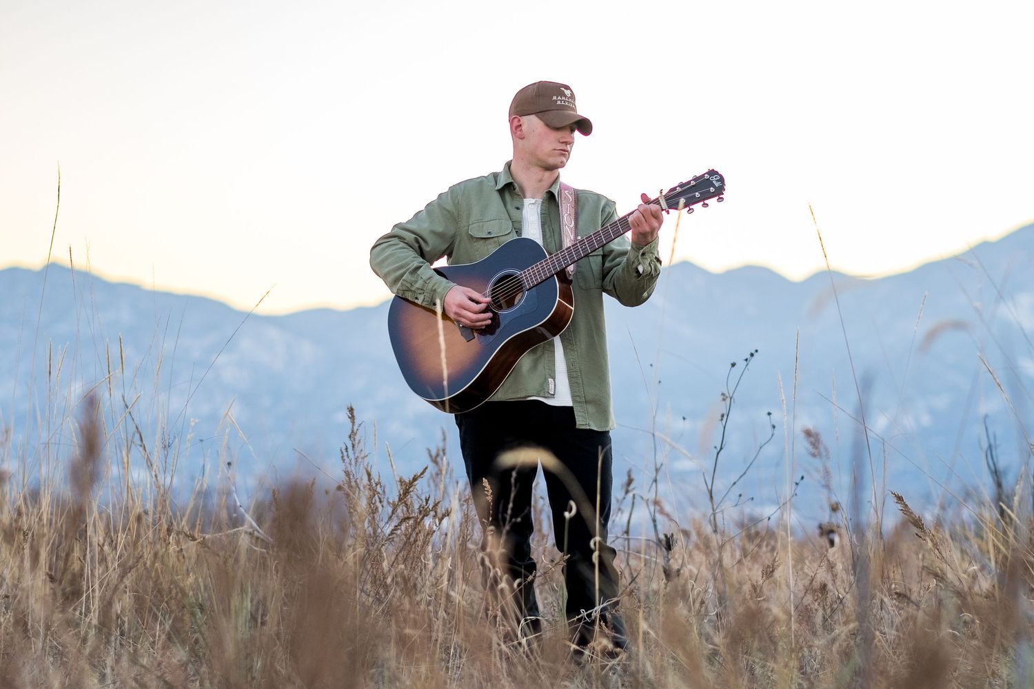 Young man strumming acoustic guitar while standing in golden field with blue mountain backdrop at sunset.