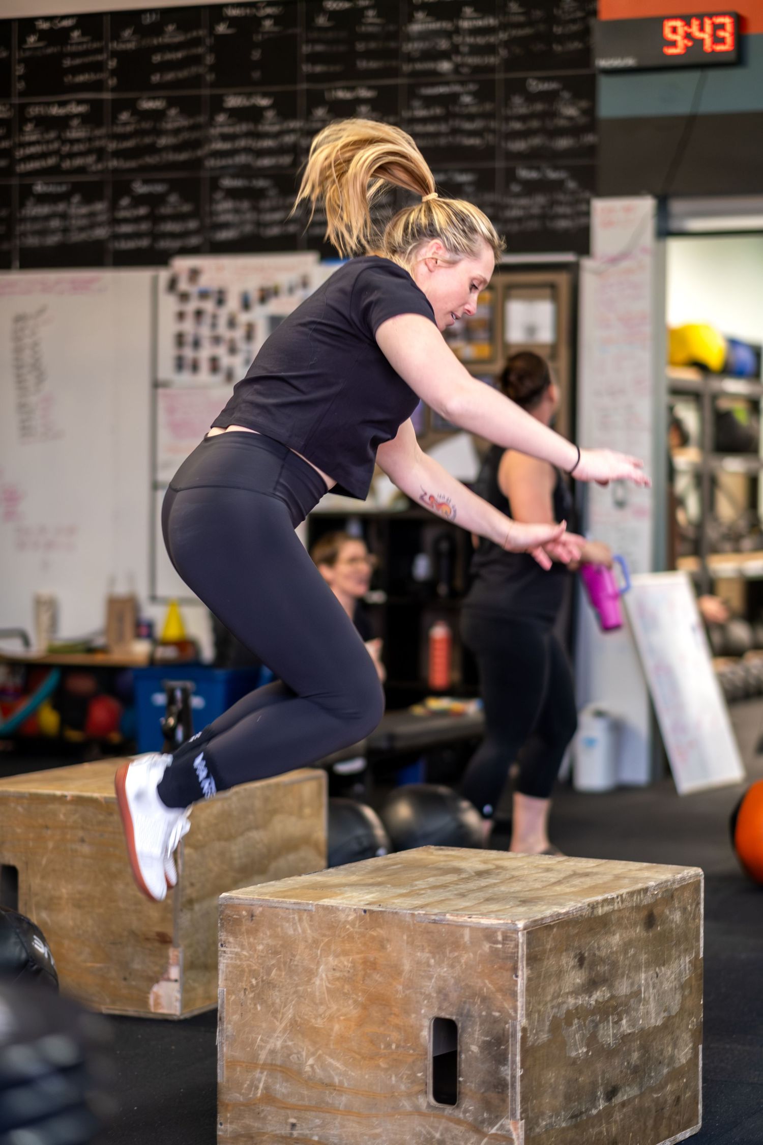 Blonde woman jumping onto a wooden plyo box during a CrossFit gym workout.