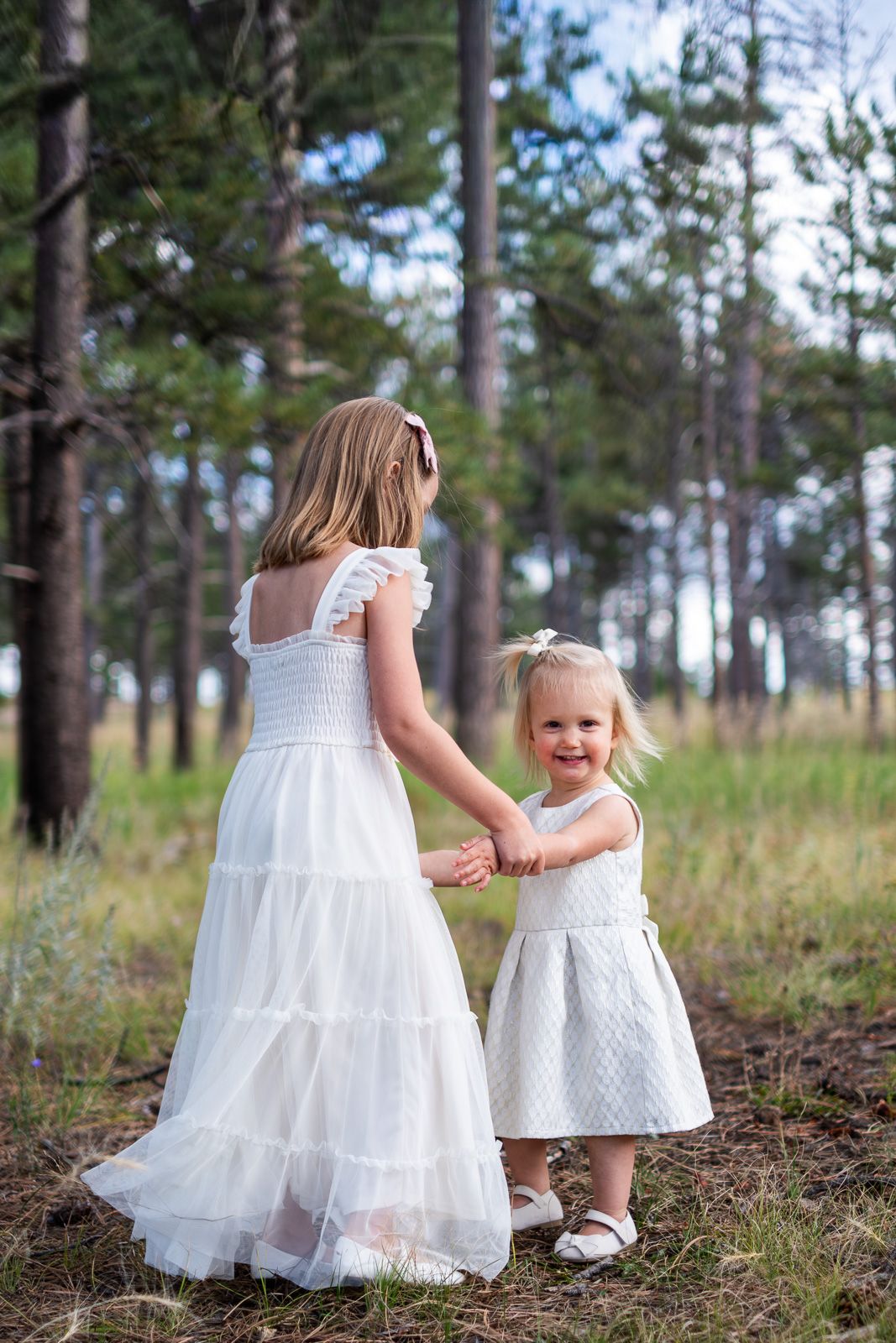 Two young sisters in white dresses hold hands while walking through a pine forest.