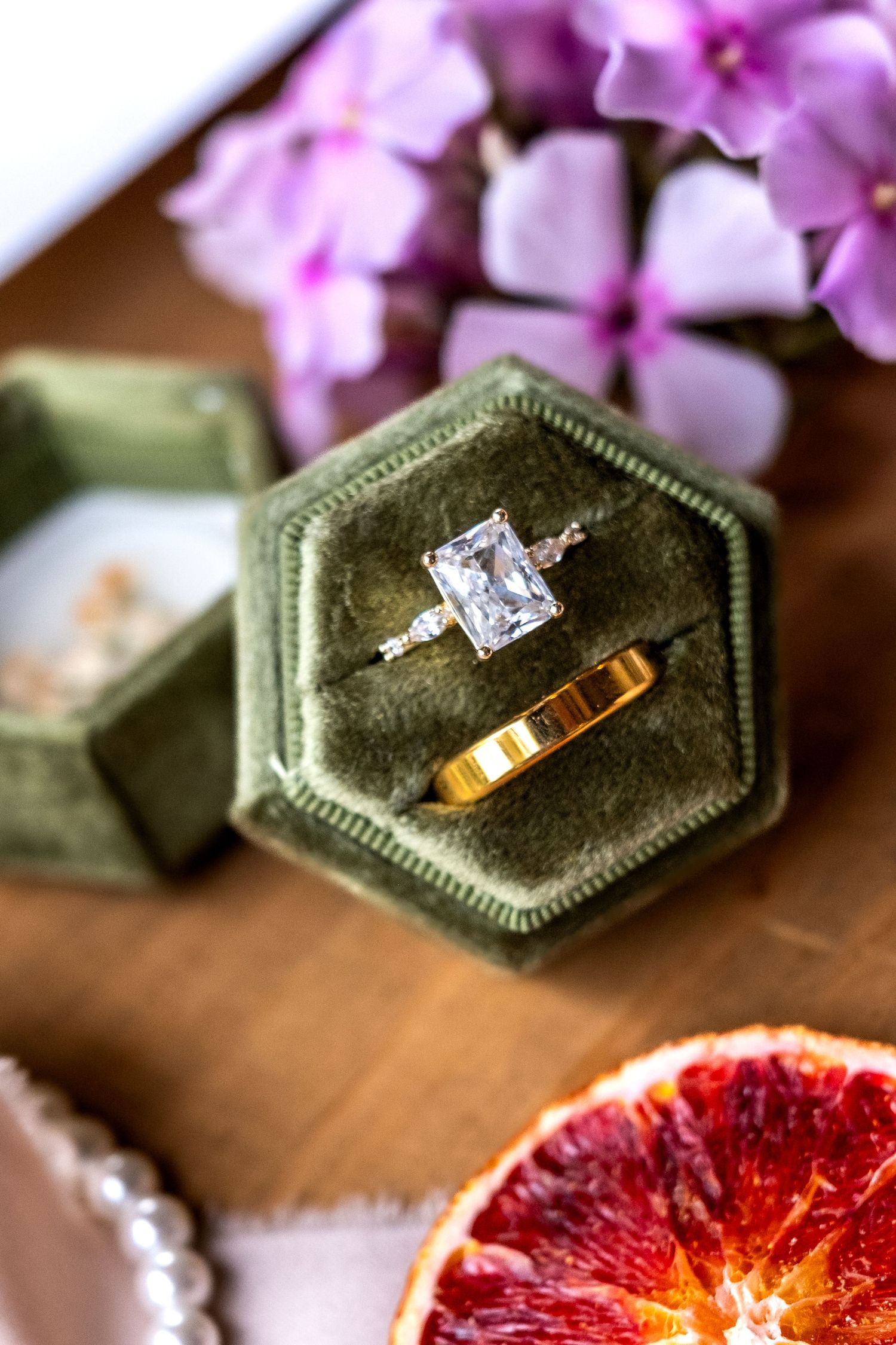 Elegant engagement ring with emerald-cut diamond in green velvet hexagon box beside gold band and pink flowers.