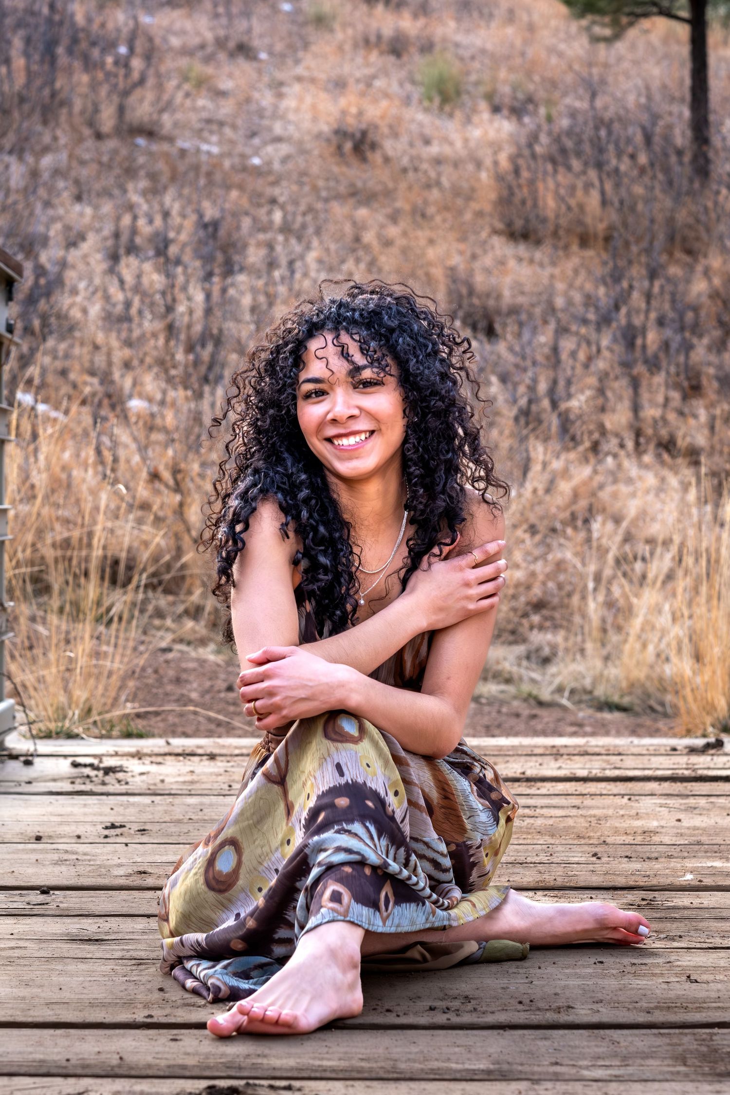 Smiling woman with curly hair sitting barefoot on wooden deck in bohemian patterned pants.