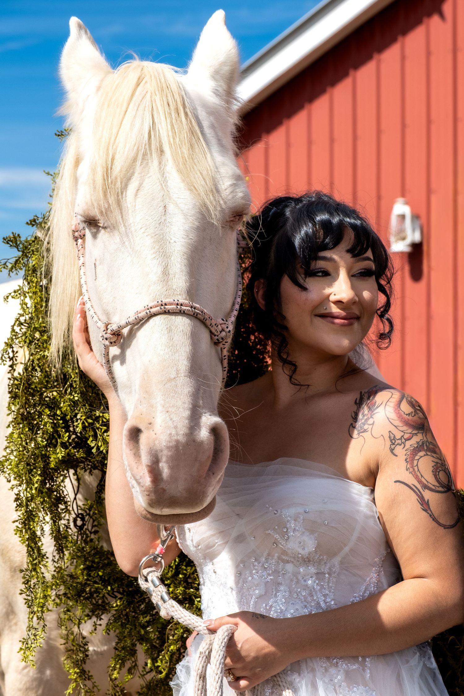 Tattooed bride in white wedding dress smiling beside a white horse in front of a red barn.