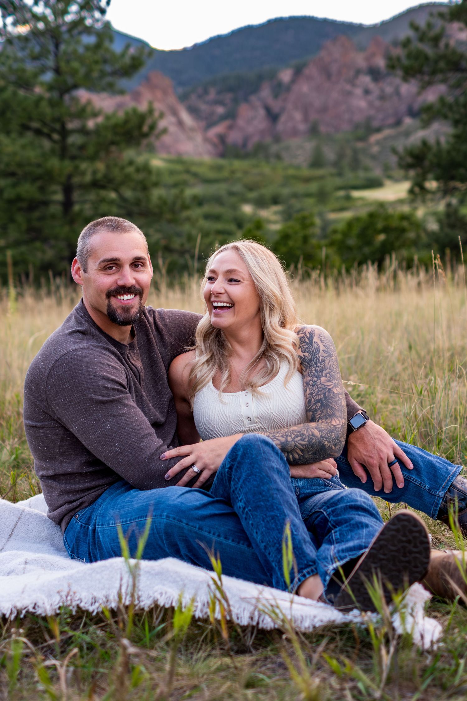 Happy couple sitting on a blanket in a mountain meadow, laughing together during an engagement session.